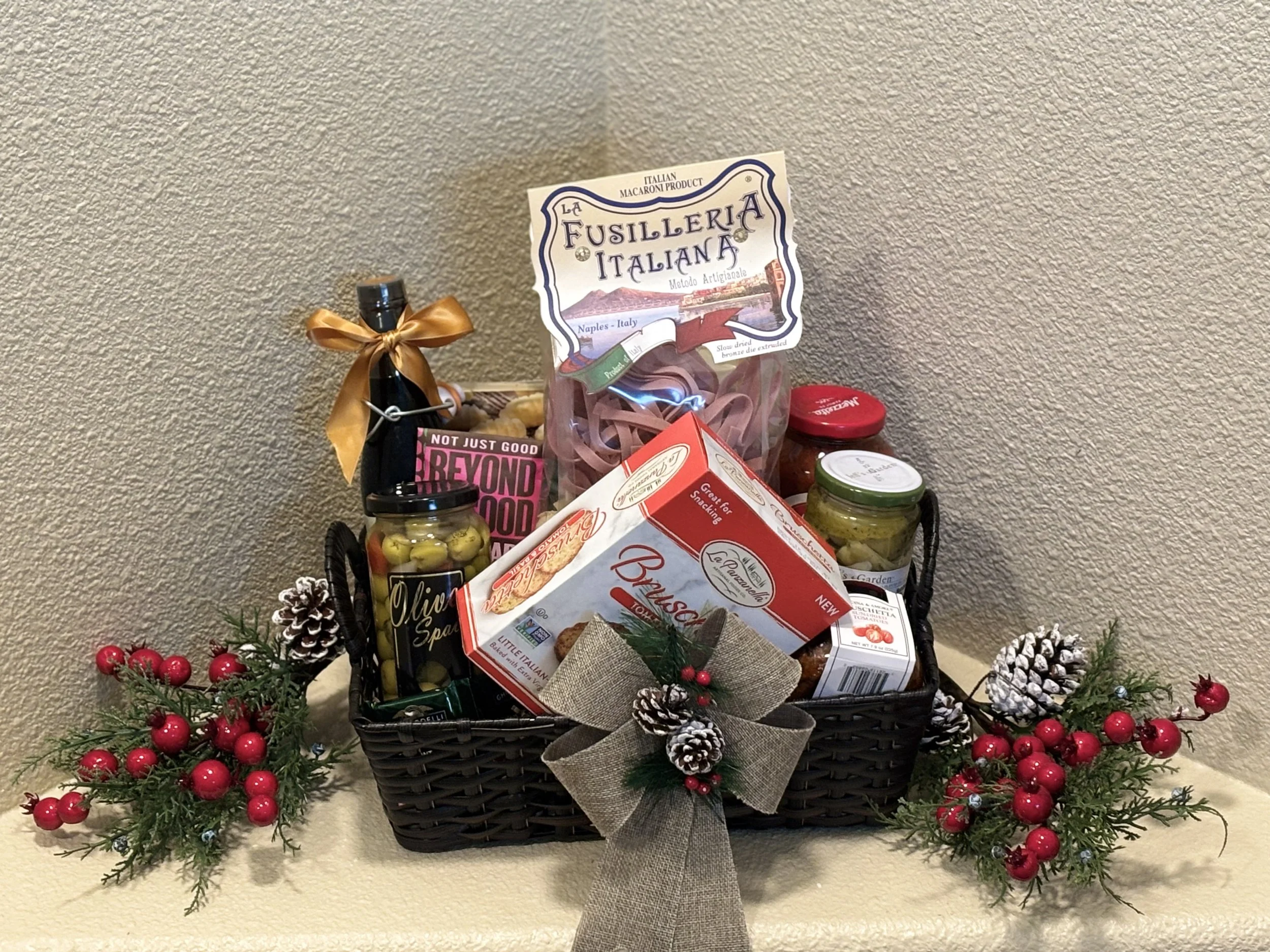 Gift basket filled with Italian food products, decorated with pinecones, holly, and a bow, placed on a beige surface against a textured wall.