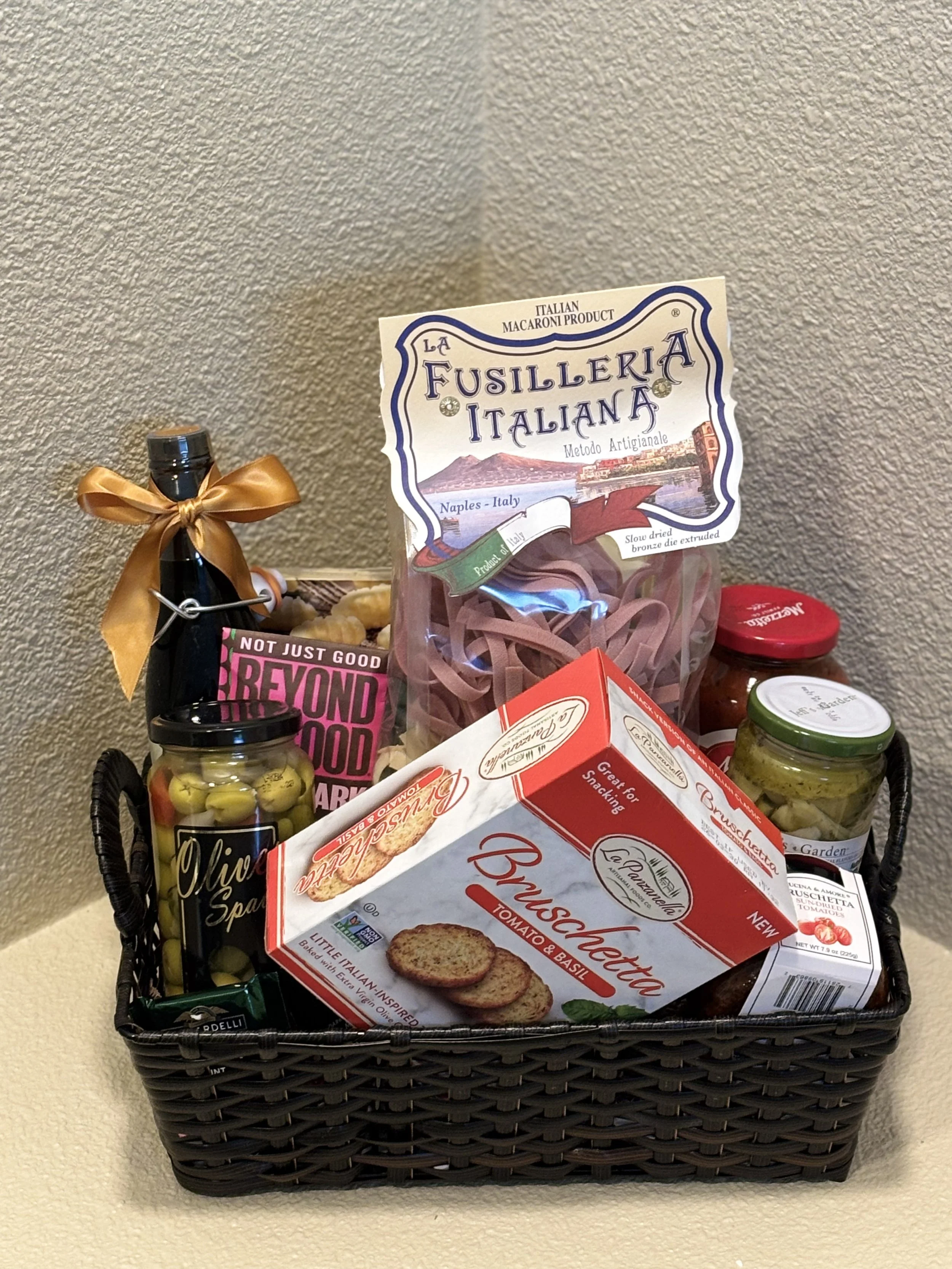 Gift basket filled with Italian food products, decorated with pinecones, holly, and a bow, placed on a beige surface against a textured wall.
