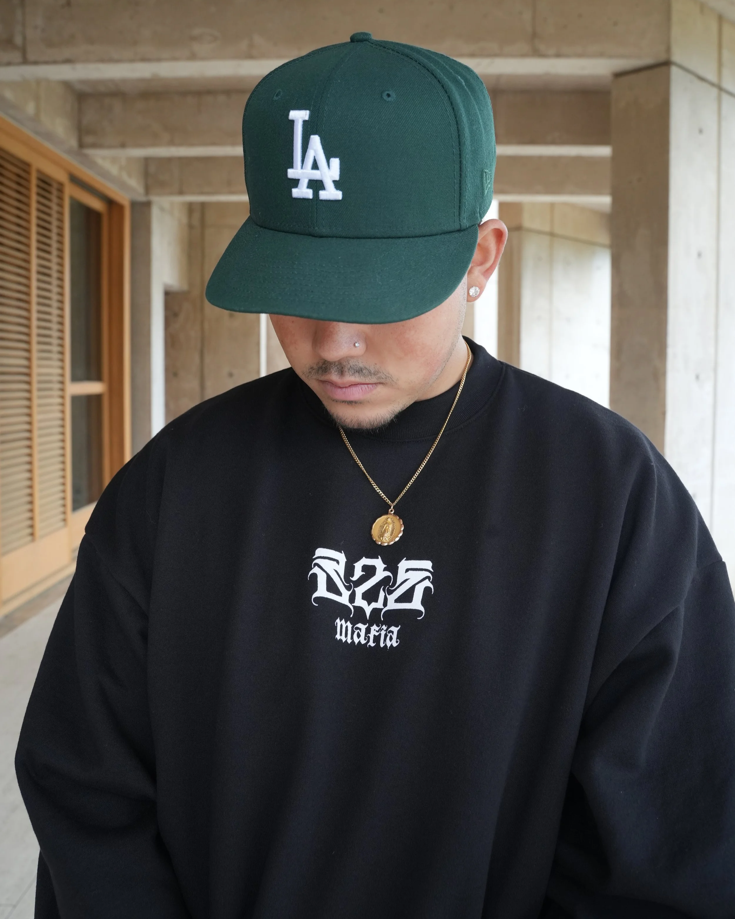 A man wearing a green Los Angeles Dodgers baseball cap, black sweatshirt with white graphic text, gold necklace with pendant, and earrings.