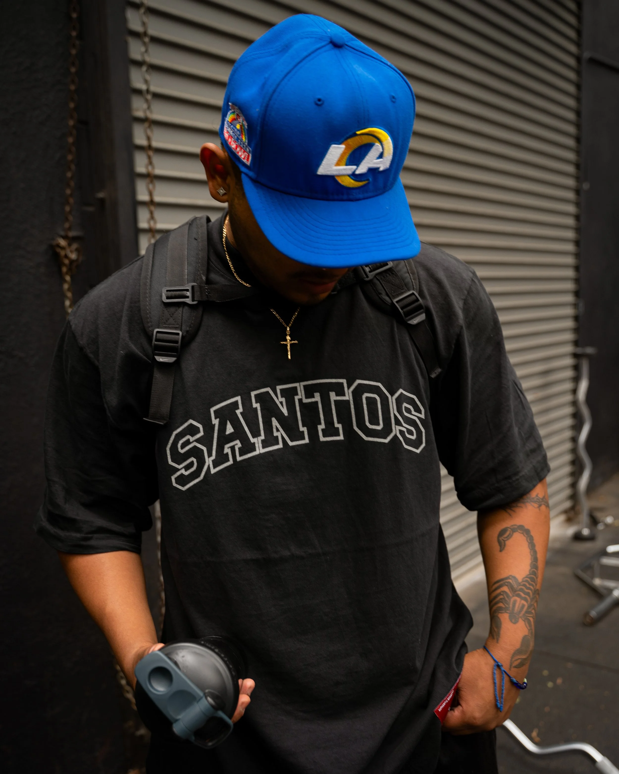 A young man wearing a blue LA Dodgers cap, a black 'SANTOS' T-shirt, gold cross necklace, and backpack, holding a bottle with a gray cap, standing in front of a closed metal roll-up door.