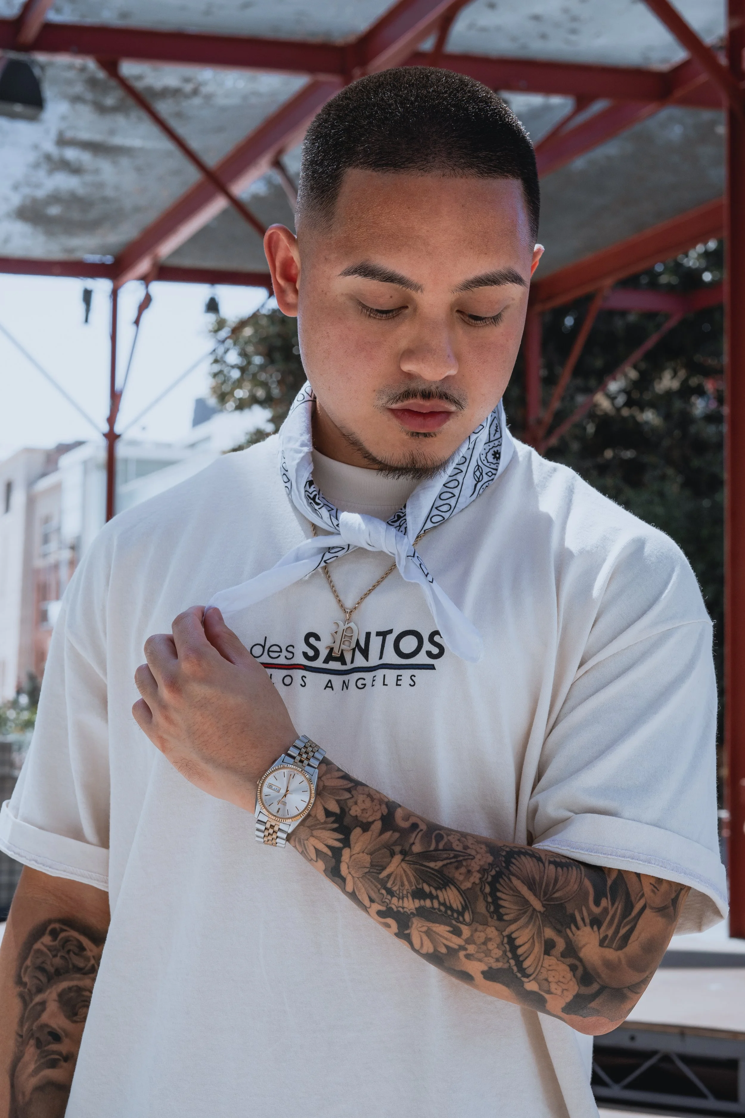 A young man with short hair and tattoos on his arm, wearing a white t-shirt, a gold watch, and a chain, standing outdoors under a metal structure, looking down and adjusting his collar.