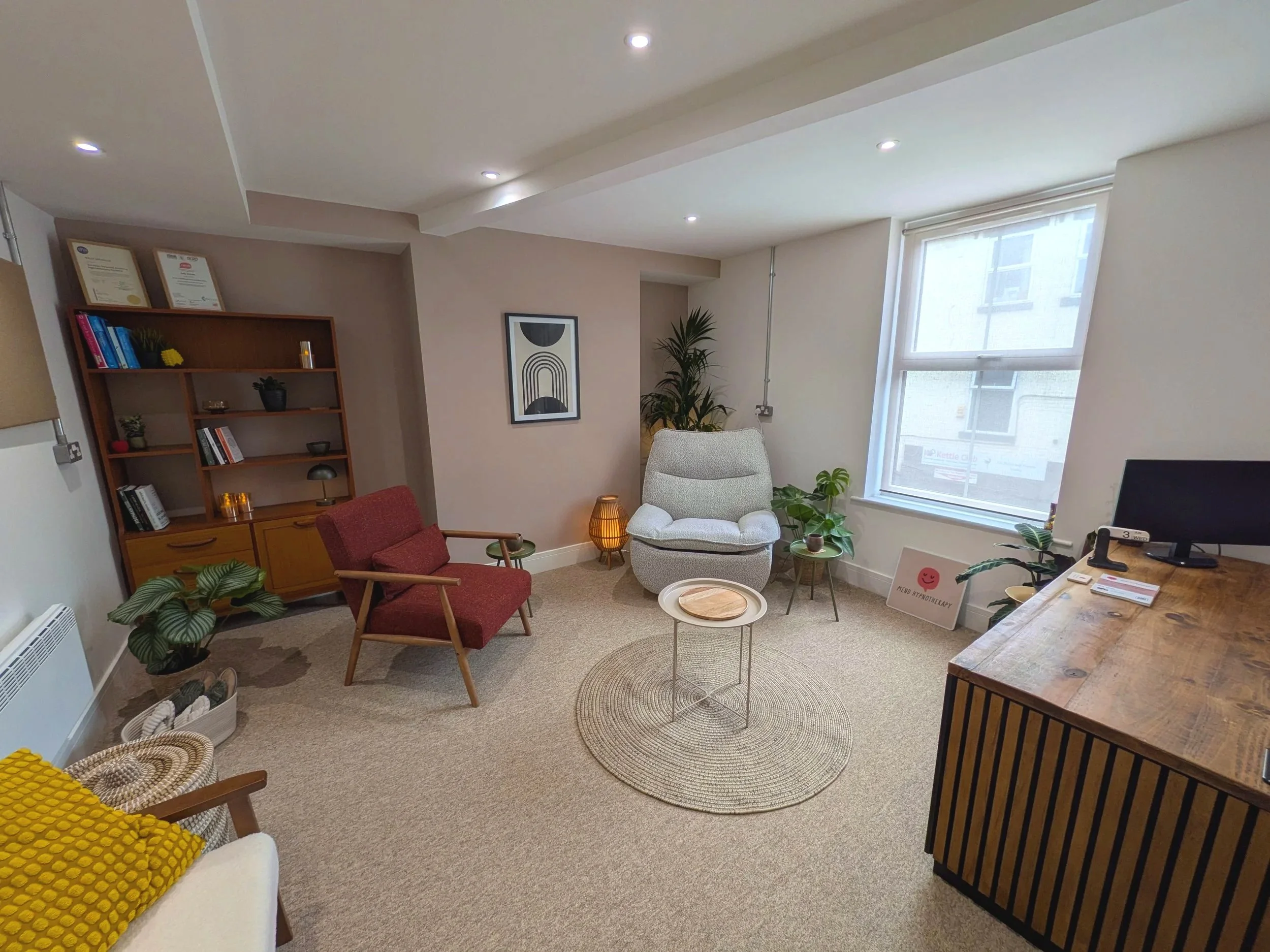 A cozy living room with a white armchair, red chair, and woven pouf, decorated with plants, modern art, and warm lighting by a large window.