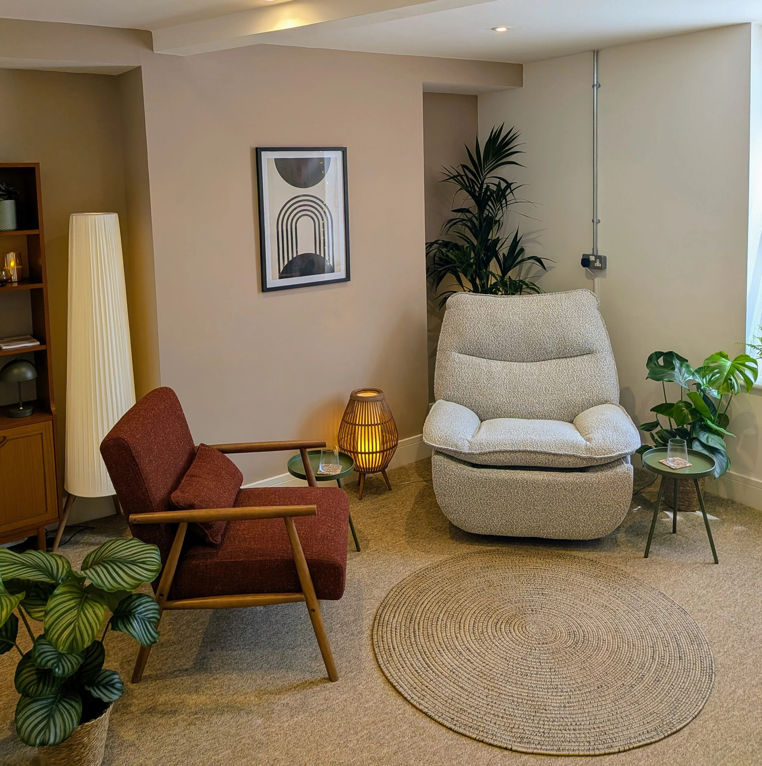 A cozy living room corner with a beige armchair and a matching beige ottoman, a potted plant, a standing lamp, a wooden shelf, and decorative wall art, with a circular woven rug on the carpeted floor.
