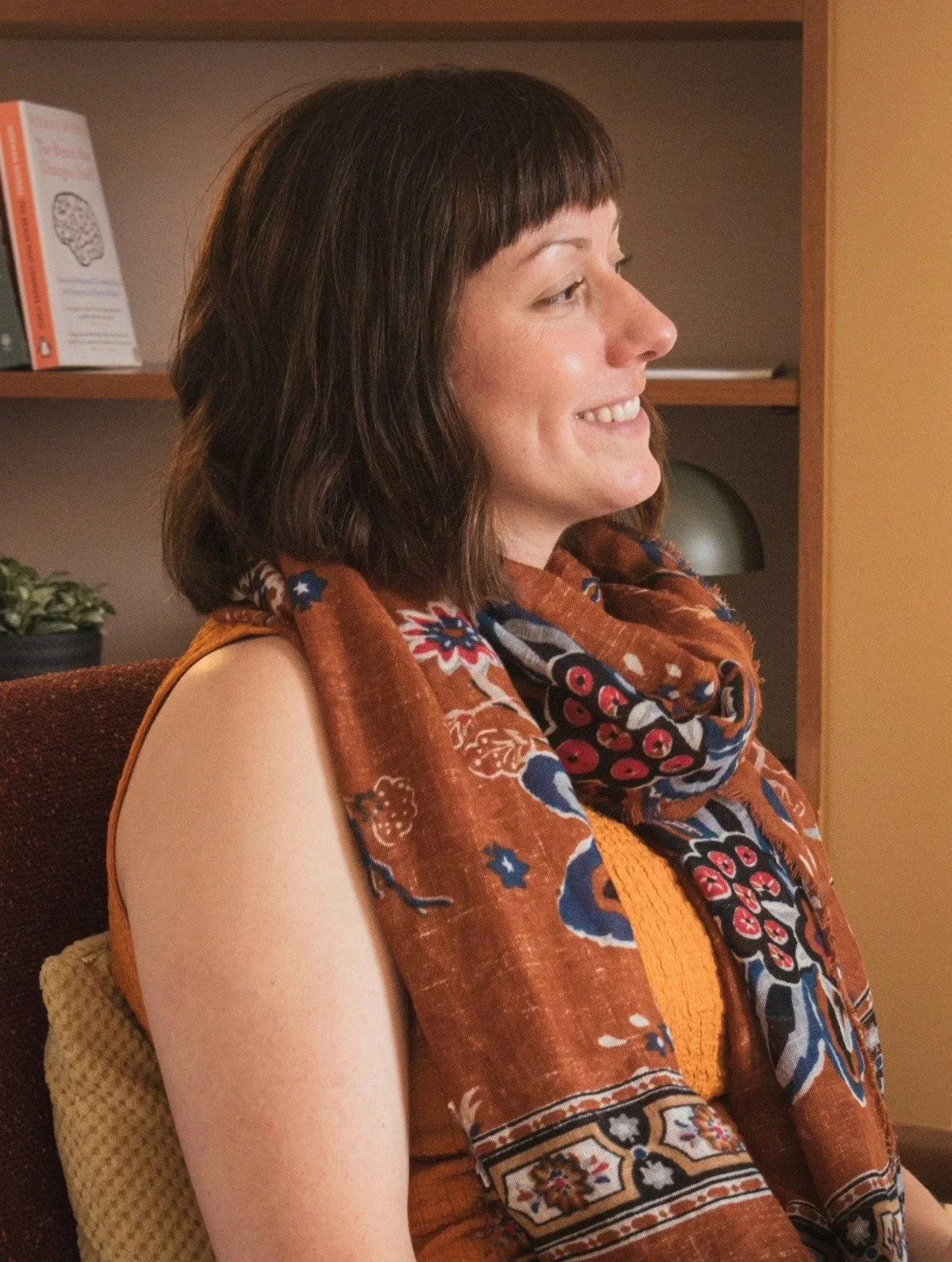 Side profile of a woman with dark brown hair, smiling, wearing a colorful patterned scarf and a sleeveless orange top, sitting on a chair in a room with shelves and decorative items in the background.