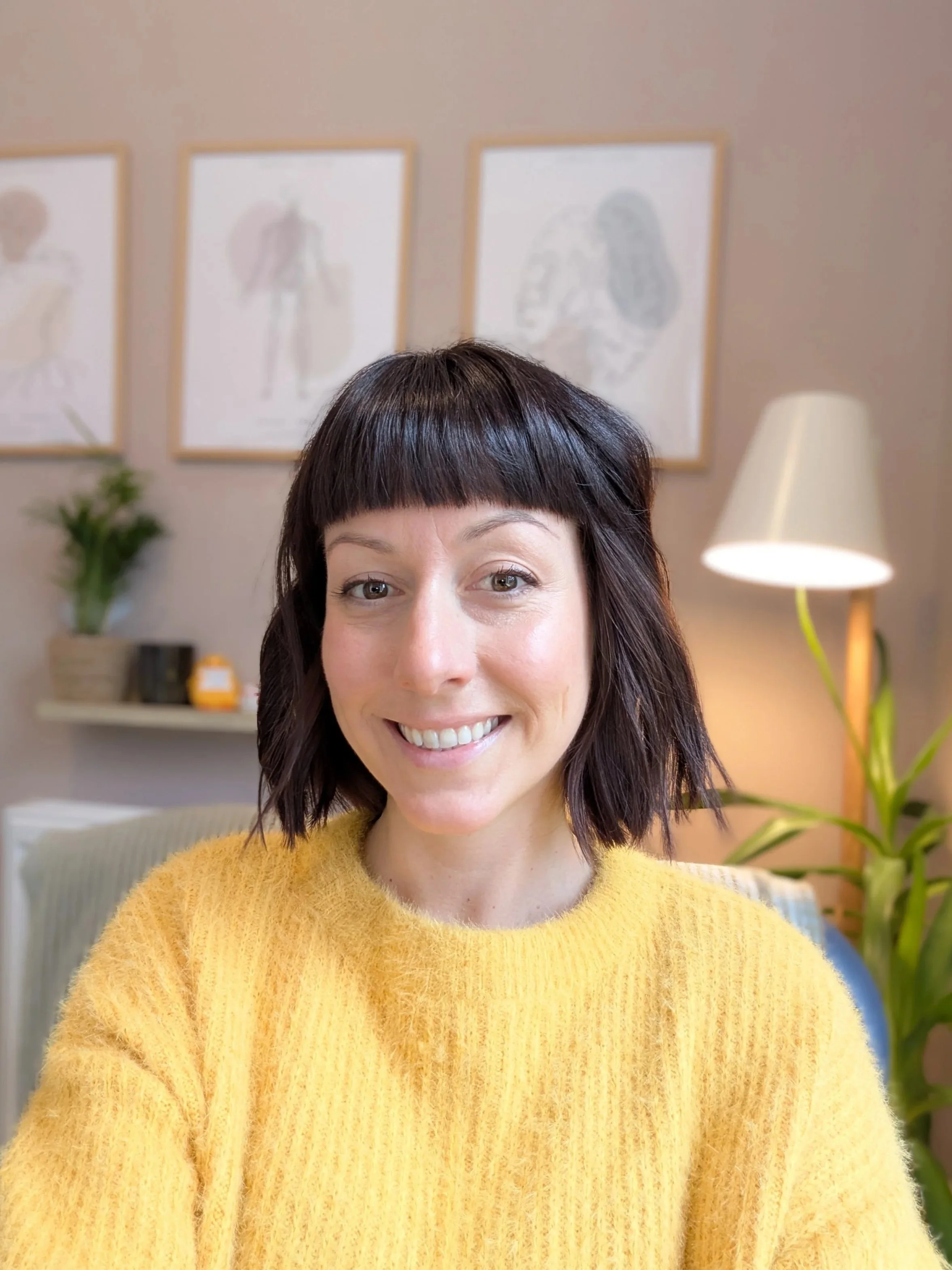 A woman with short dark hair and bangs, smiling and wearing a yellow sweater, sitting in a well-lit room with framed artwork on the wall and plants in the background.