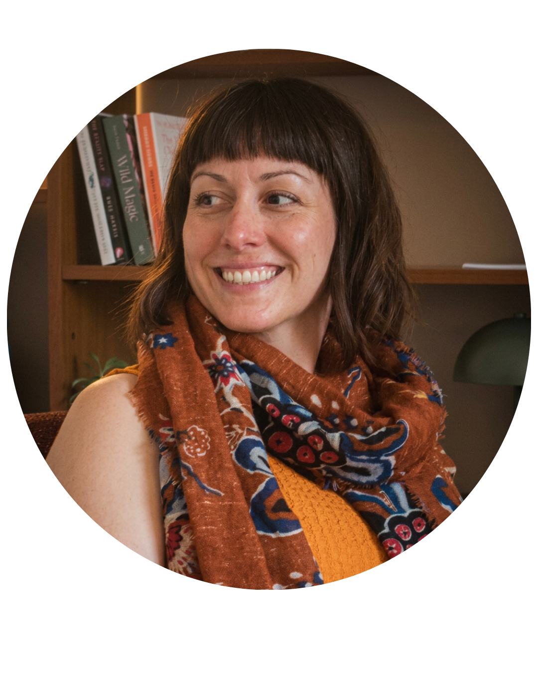 A woman with shoulder-length brown hair and a patterned scarf smiling inside a room with bookshelves.