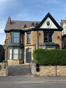 A Victorian-style house with brick exterior, bay windows, and a gabled roof, with a green hedge and stone wall in front under a clear blue sky.