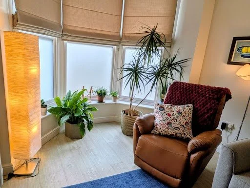 Corner of a cozy living room with large windows, potted plants, a brown recliner with a decorative pillow, a gray armchair, and a tall floor lamp.