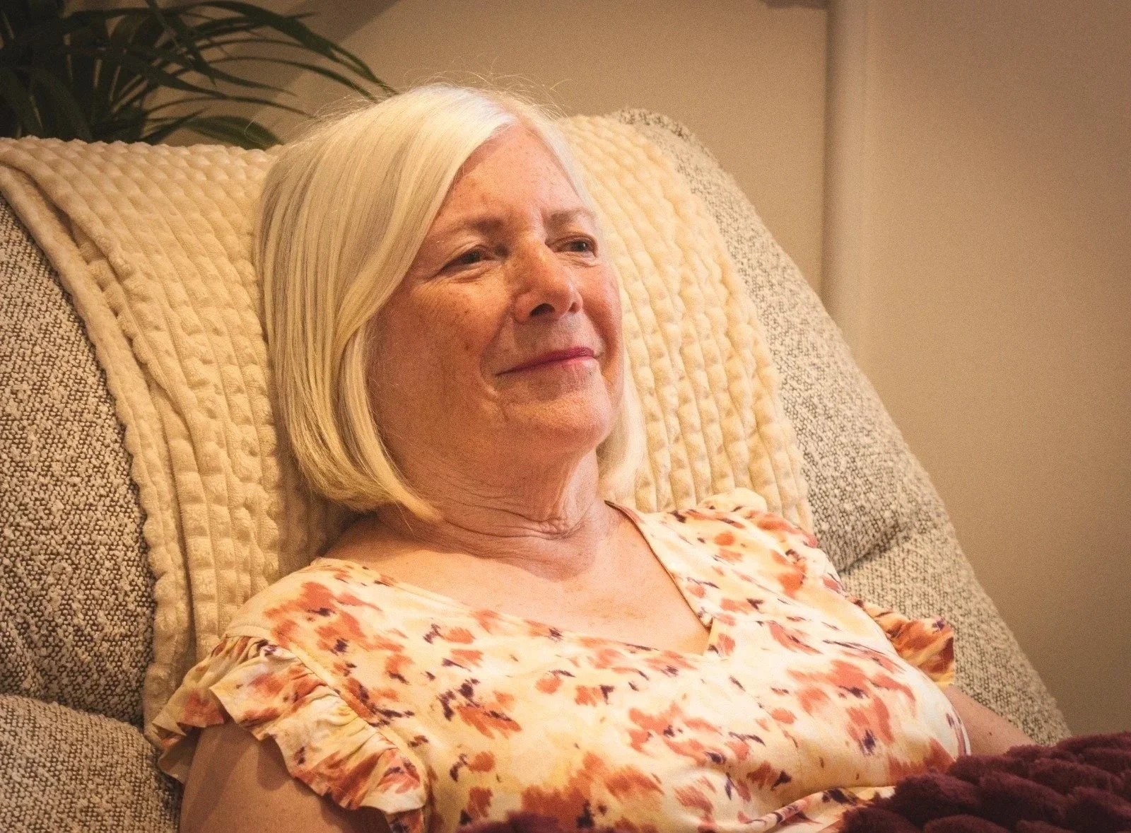 An older woman with blonde hair sitting on a beige couch, smiling gently, wearing a floral top, in a cozy indoor setting.