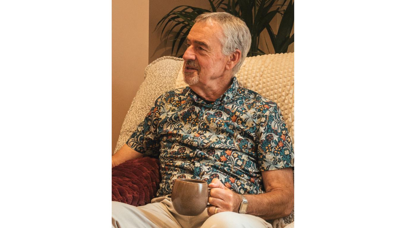 An elderly man with gray hair and a beard sitting on a beige textured armchair, wearing a colorful floral shirt, holding a brown mug, and looking to his left.