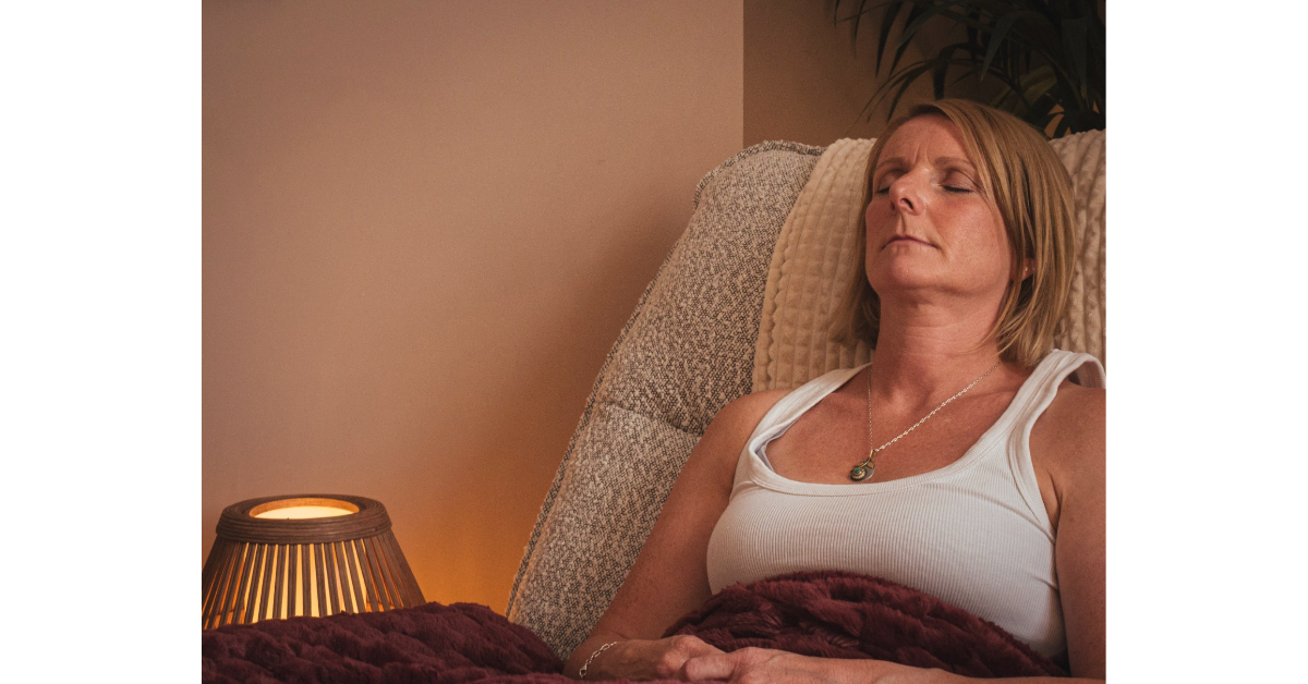 A woman resting with her eyes closed, sitting in bed under a maroon blanket, with a beige pillow behind her head and a bedside lamp on her left, in a softly lit room.