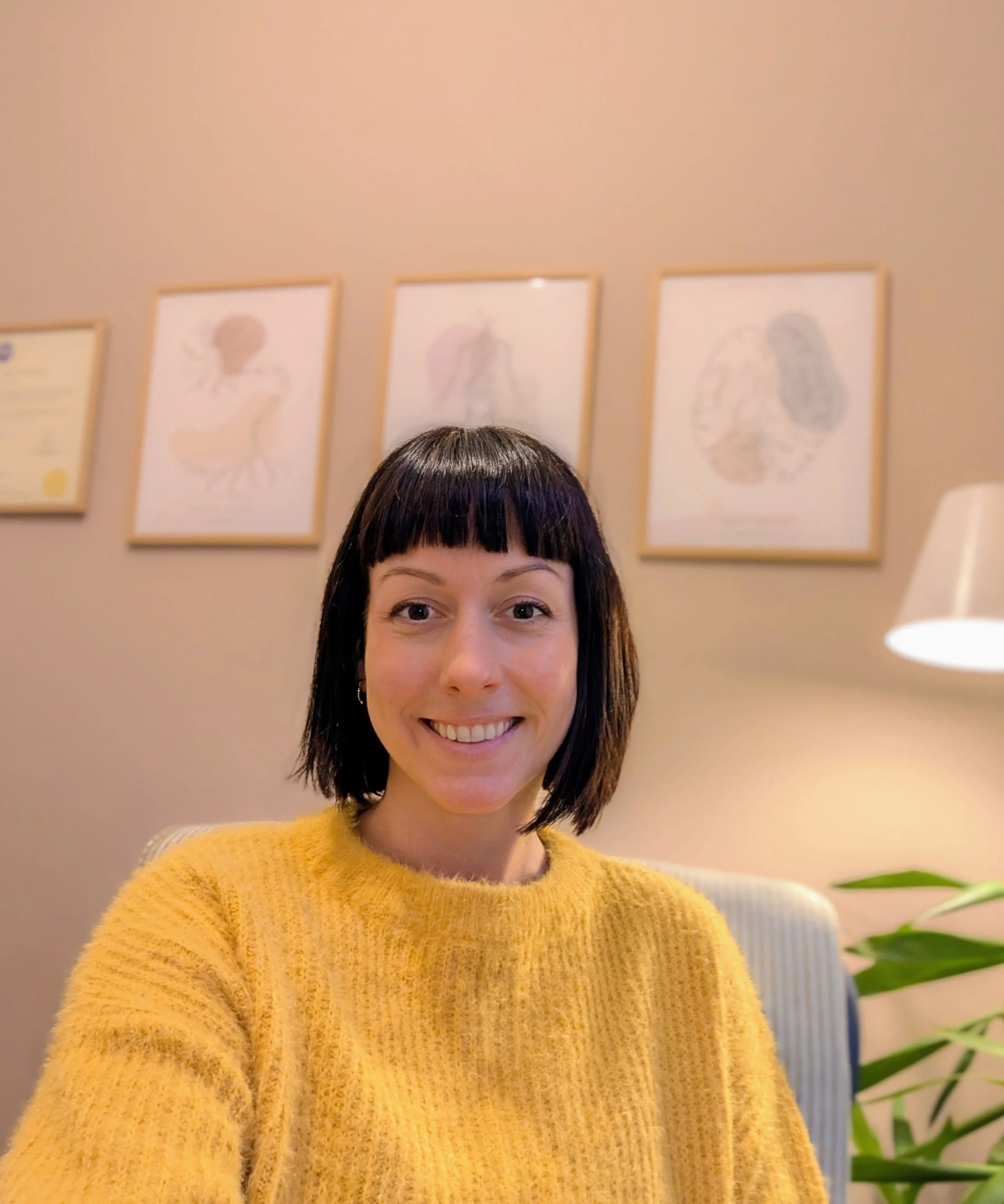 A woman with short dark hair, wearing a yellow sweater, smiling at the camera in a room with beige walls and framed prints of anatomical diagrams in the background.