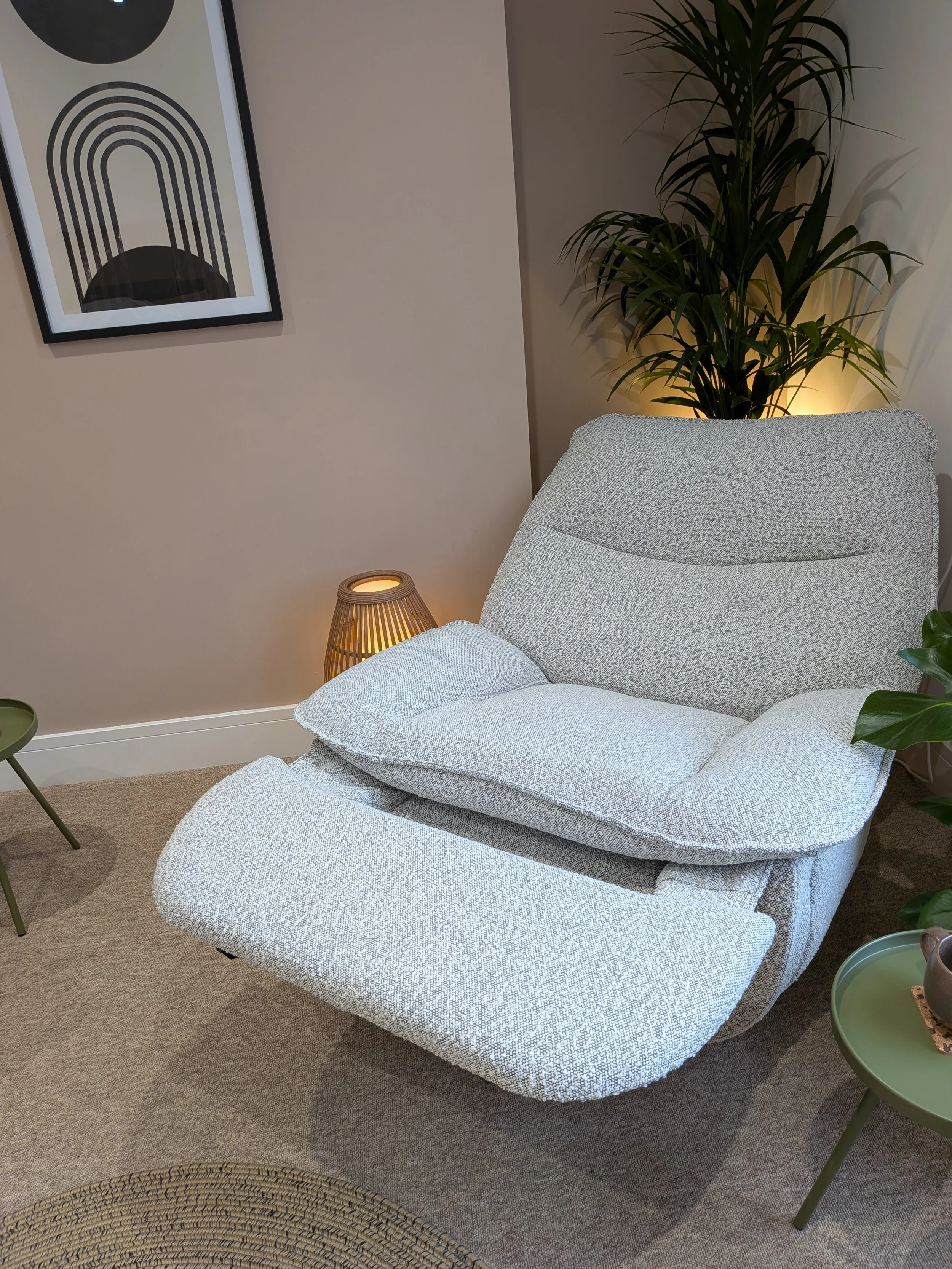 A cozy living room corner with a white textured chaise lounge, a green side table, a woven round rug, a floor lamp, and a large home plant near a beige wall with a framed abstract art print.