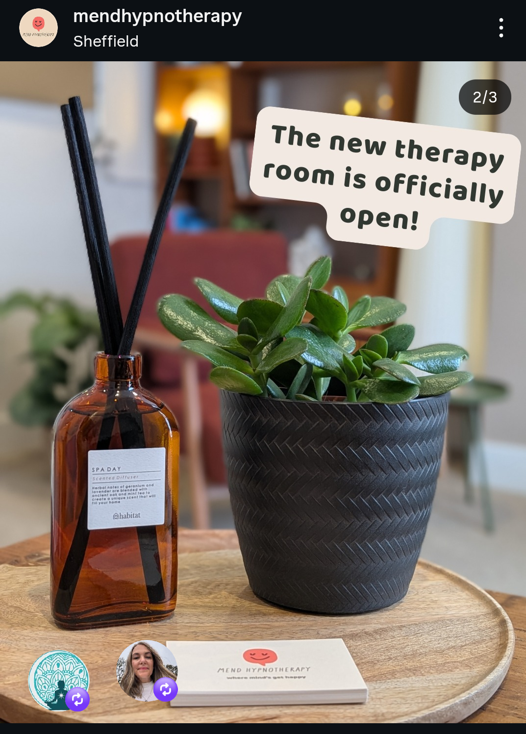 A plant in a black textured pot and a brown glass diffuser bottle with black reed sticks on a wooden tray, with a sign saying 'The new therapy room is officially open' and a card from Mend Hypnotherapy on the tray.