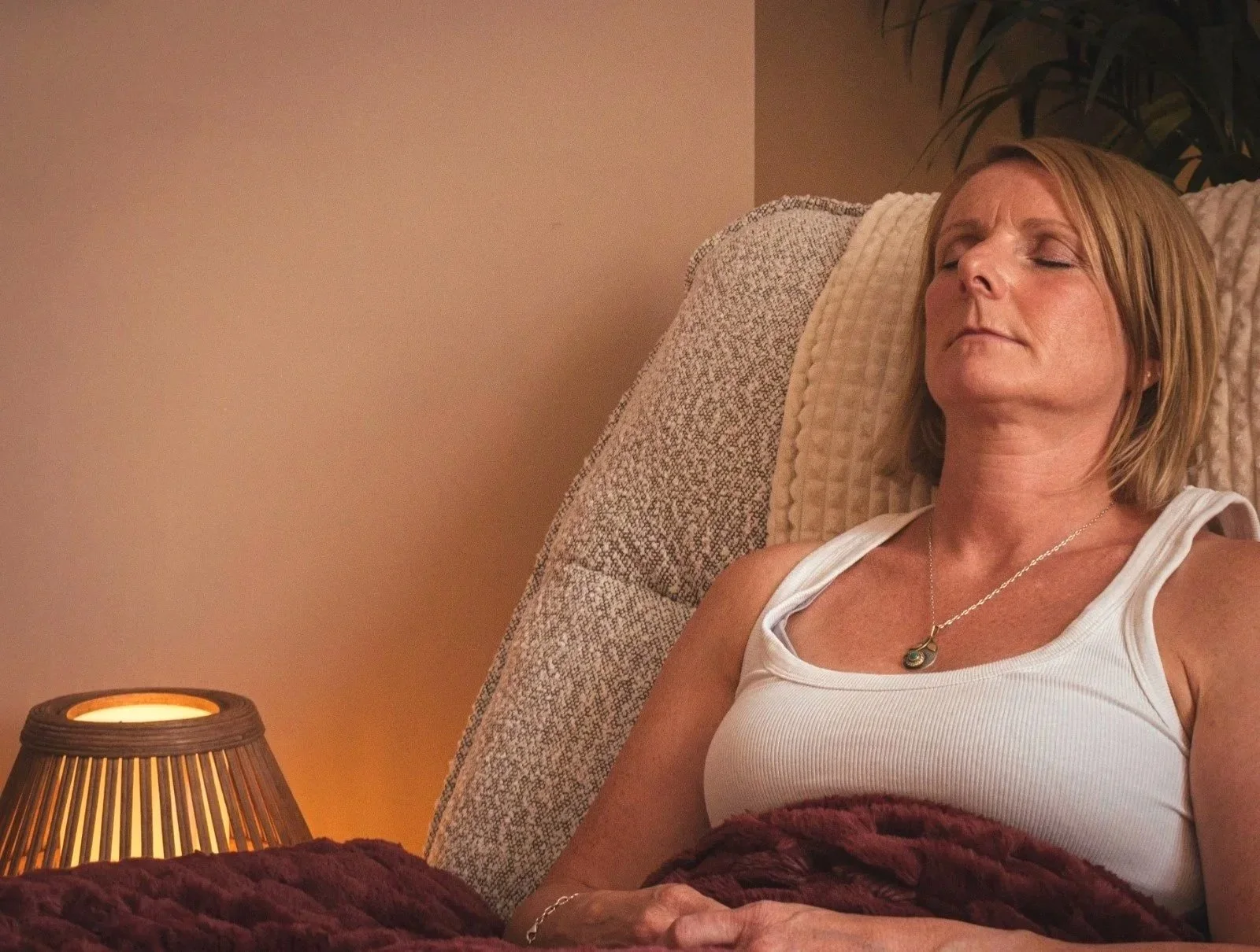 A woman with short blonde hair resting with eyes closed on a beige pillow, wearing a white sleeveless top and a necklace, sitting in a cozy room with a warm-colored wall and a lamp.