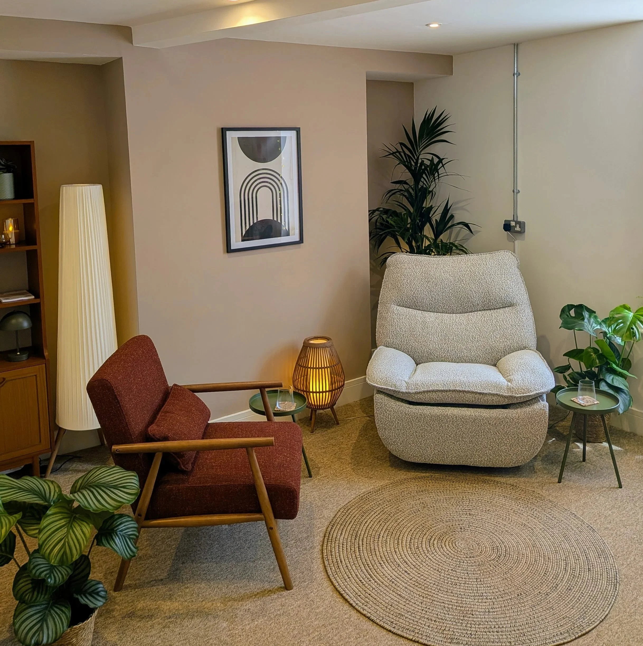 A cozy living room corner with a white armchair, a small round green side table with a drink, a tall floor lamp with a striped shade, a large patterned rug, a tall potted plant, framed abstract art on the beige wall, a red armchair partly visible, and a wooden bookshelf in the background.