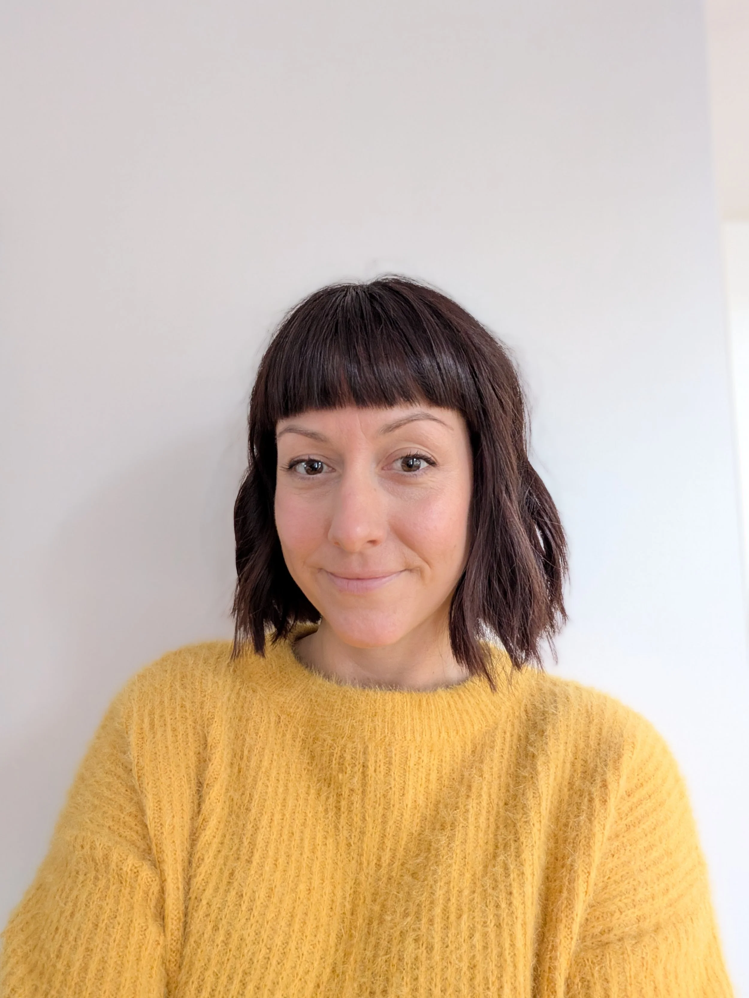 A woman with short, wavy dark hair and bangs wearing a yellow sweater, smiling at the camera against a plain white wall.