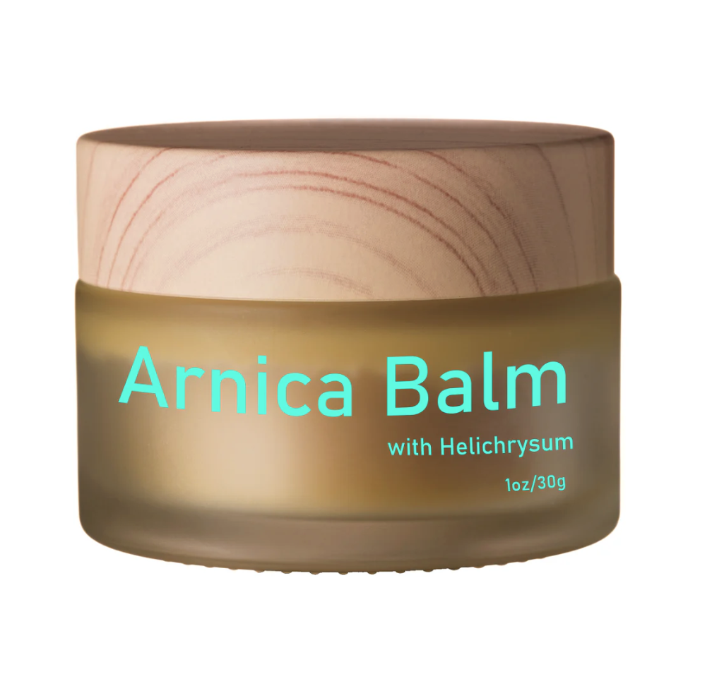 A jar of Arnica Balm with a wooden lid, labeled with teal text that says 'Arnica Balm with Helichrysum 1oz/30g'.
