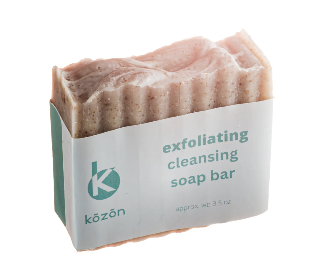 A bar of exfoliating cleansing soap with a pink, textured surface in a white and teal wrapper labeled 'exfoliating cleansing soap bar' and the brand name 'kōzōn'