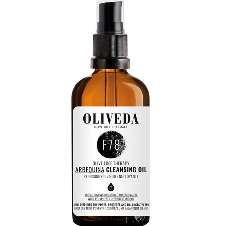 Amber glass spray bottle labeled Oliveda Arbequina Cleansing Oil with black spray nozzle and white label.