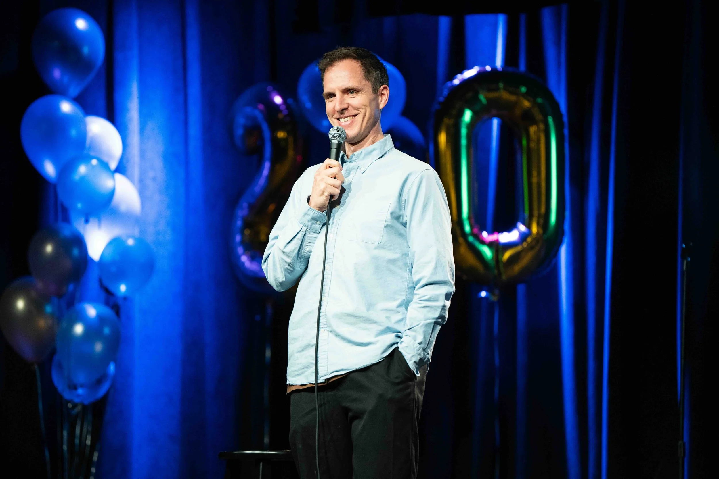 A man performing stand-up comedy on stage with blue balloons and large gold balloons spelling the number 20 in the background.