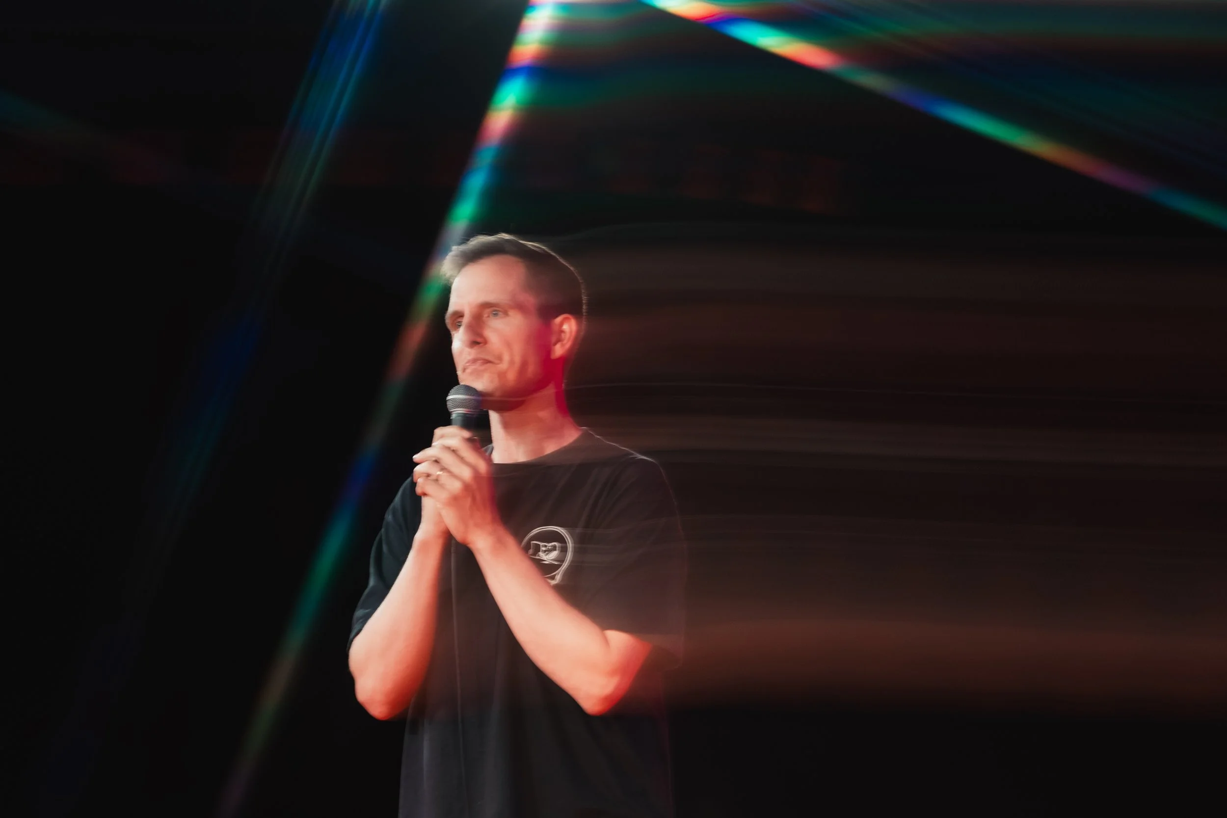 A man holding a microphone standing on a stage with colorful lights creating streaks in the background.