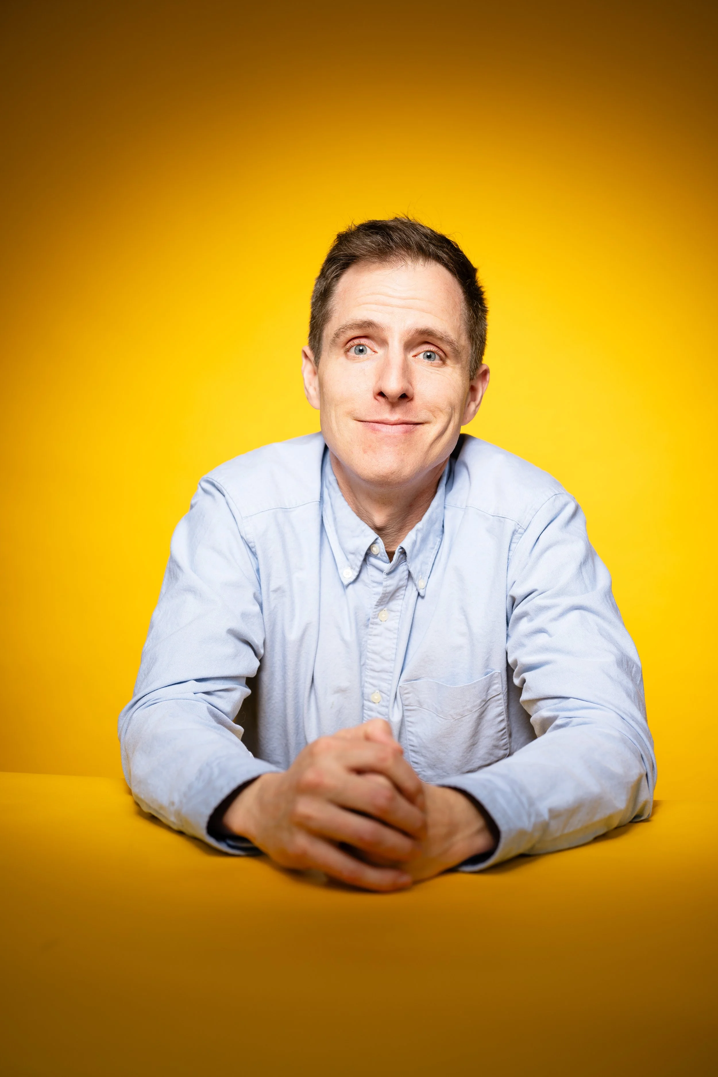 A man with short dark hair, wearing a light blue button-up shirt, sitting at a yellow table against a yellow background, smiling with hands clasped.
