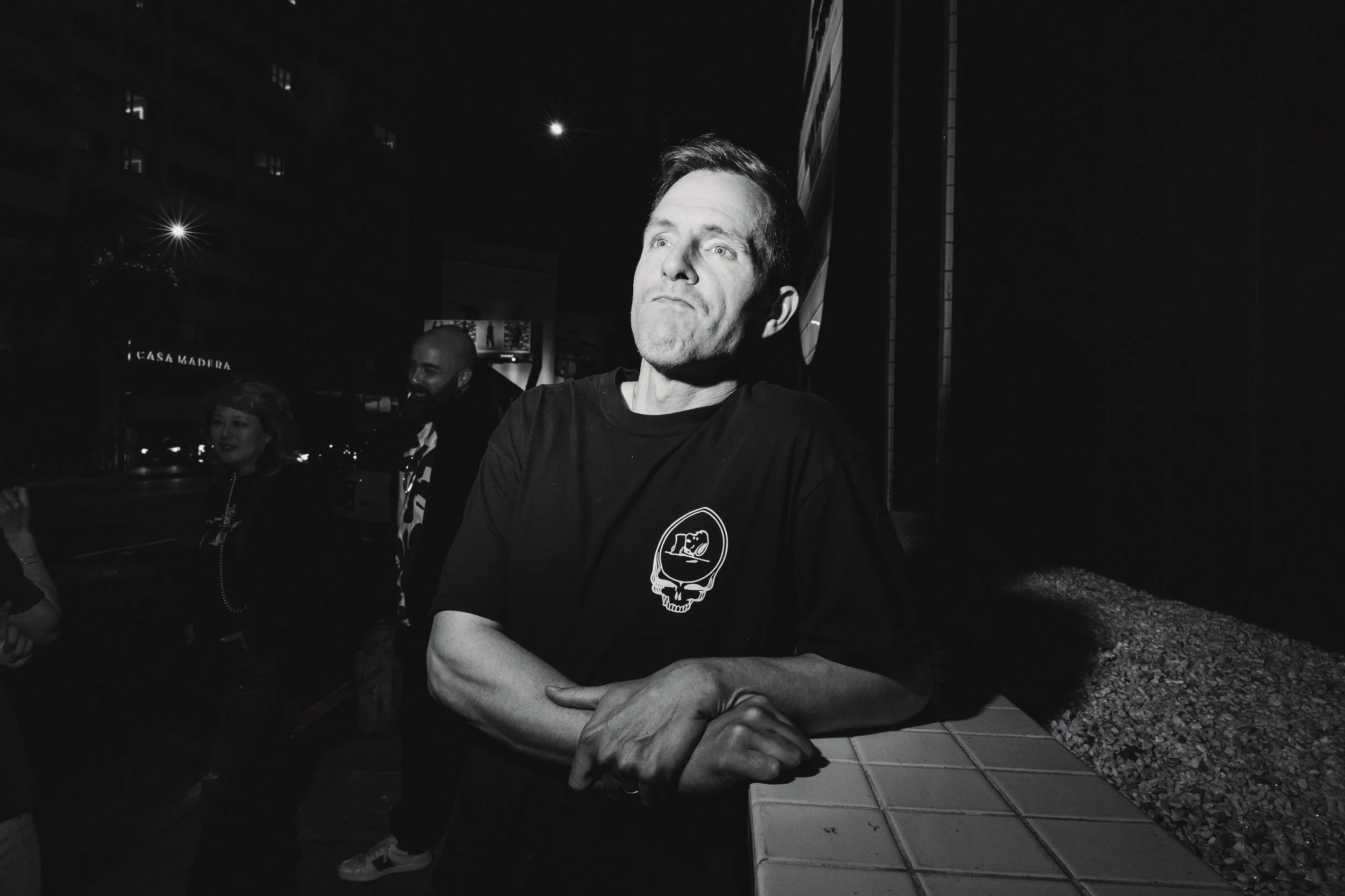 Black and white photo of a man with short hair and a serious expression, leaning on a tiled ledge with his arms crossed, wearing a dark t-shirt with a skull graphic. In the background, there are three other people and a building at night.