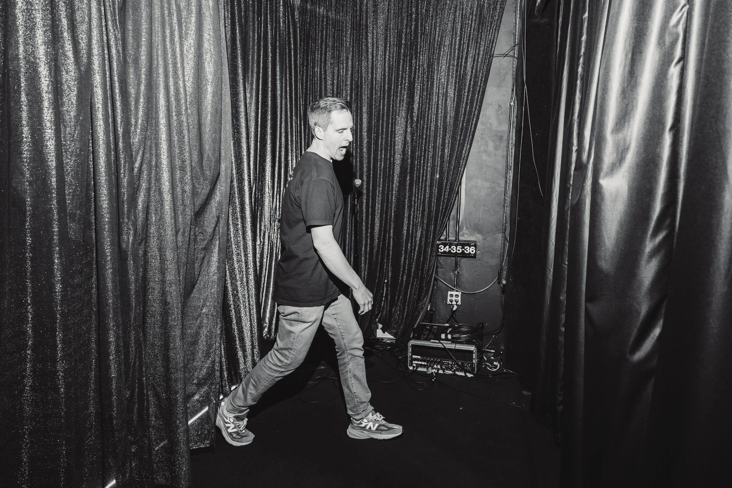A man walking in a backstage area with black curtains on either side, near stage equipment and a digital timer display.