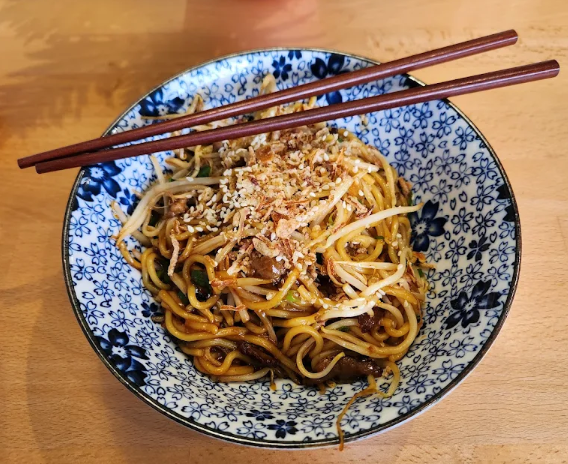 Bowl of Asian-style stir-fried noodles with vegetables, topped with sesame seeds, served on a wooden table with chopsticks resting on the bowl.