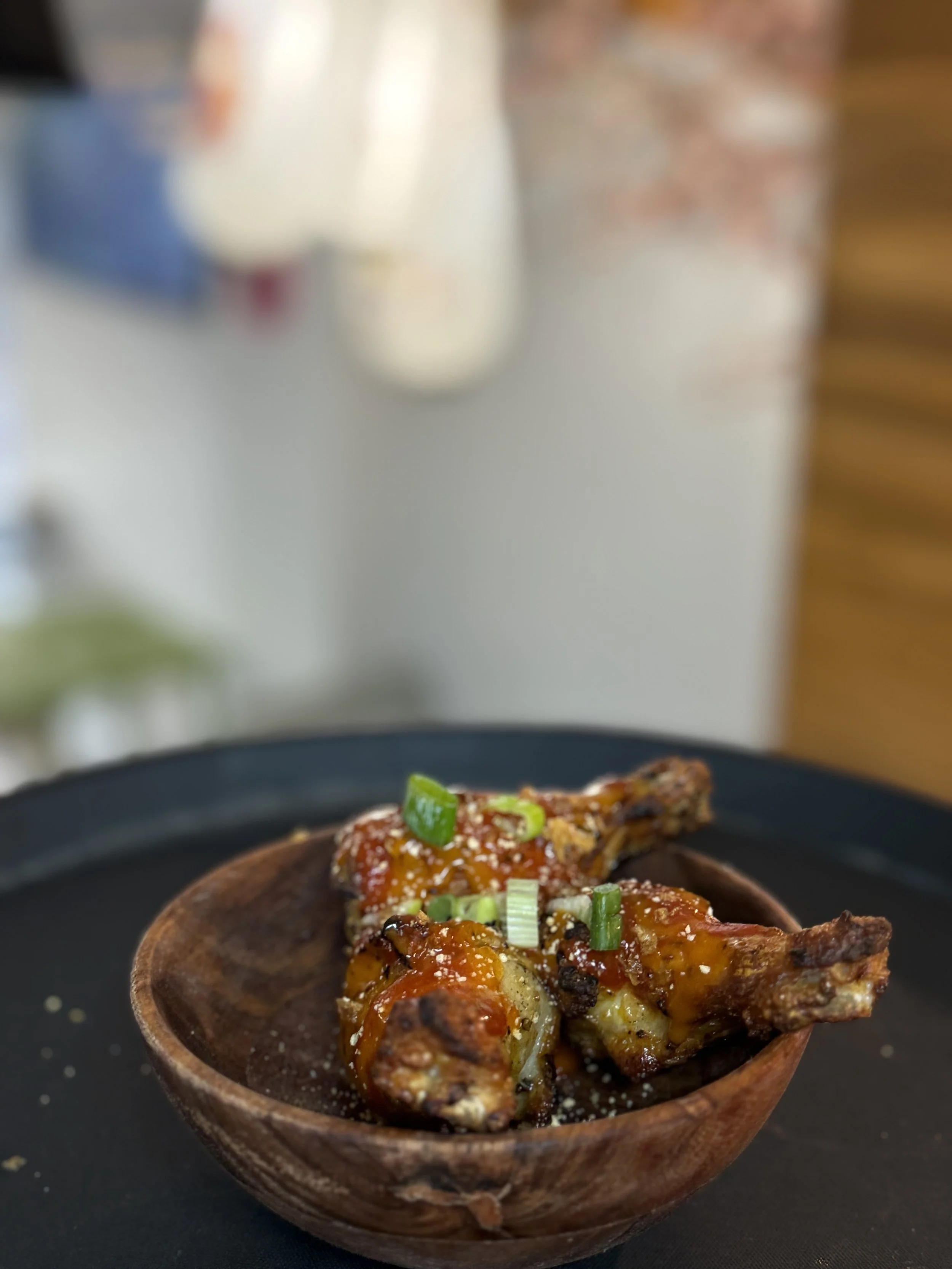 Close-up of two glazed chicken drumsticks garnished with chopped green onions served in a wooden bowl on a black tray.