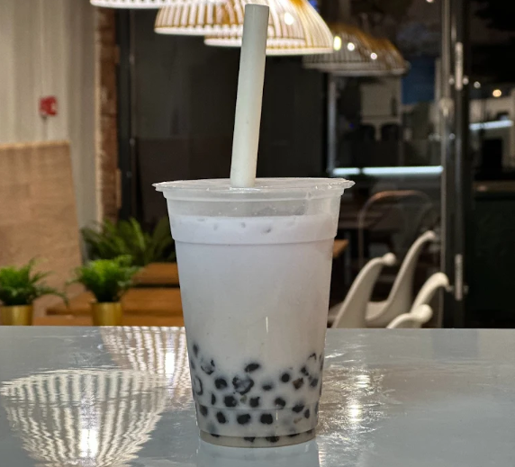 A cup of bubble tea with black tapioca pearls at the bottom, a wide straw, and a plastic lid, on a reflective table inside a cafe.
