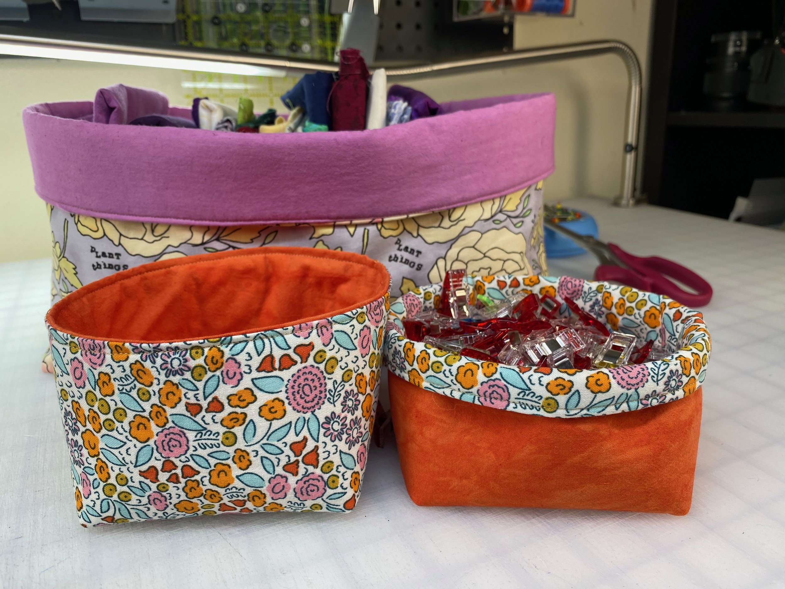 Two fabric baskets filled with sewing supplies on a white surface, with a pink container on top containing fabric and small craft items, and another patterned basket below filled with clear plastic clips, scissors, and fabric pieces.