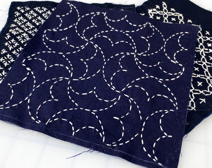 Three dark blue fabric pieces with white embroidered patterns, including curved dashed lines and geometric designs.