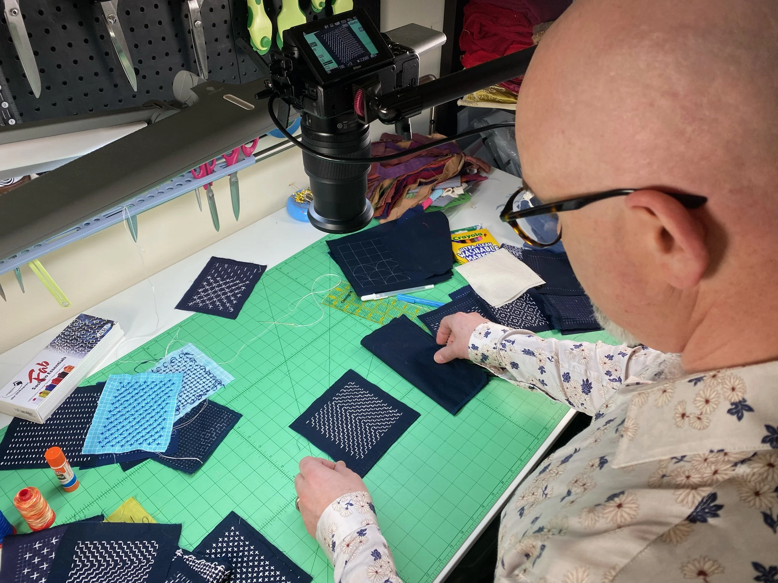 A person with glasses and a floral shirt is working at a craft table with various fabric samples, thread, and sewing supplies. The person is holding a dark fabric piece on a green cutting mat.