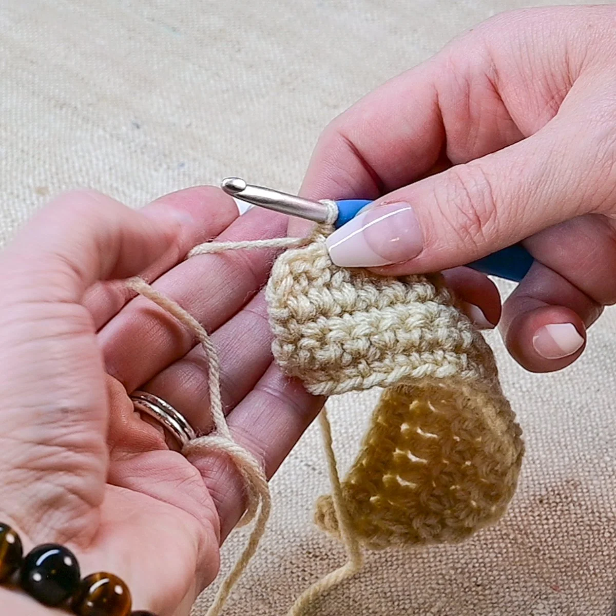 Hands crocheting beige yarn into a small project with a crochet hook, on a neutral fabric surface.