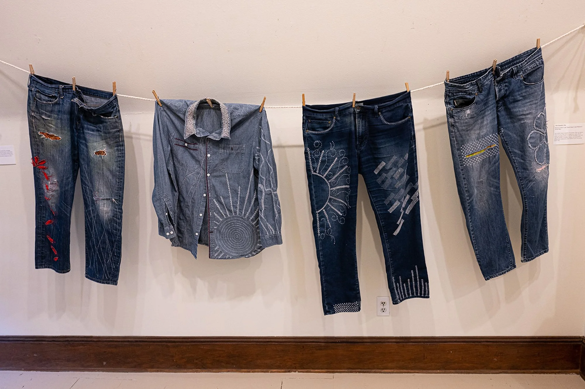 Four pairs of jeans and one denim shirt hanging on a clothesline with clothespins, displayed in an exhibition.