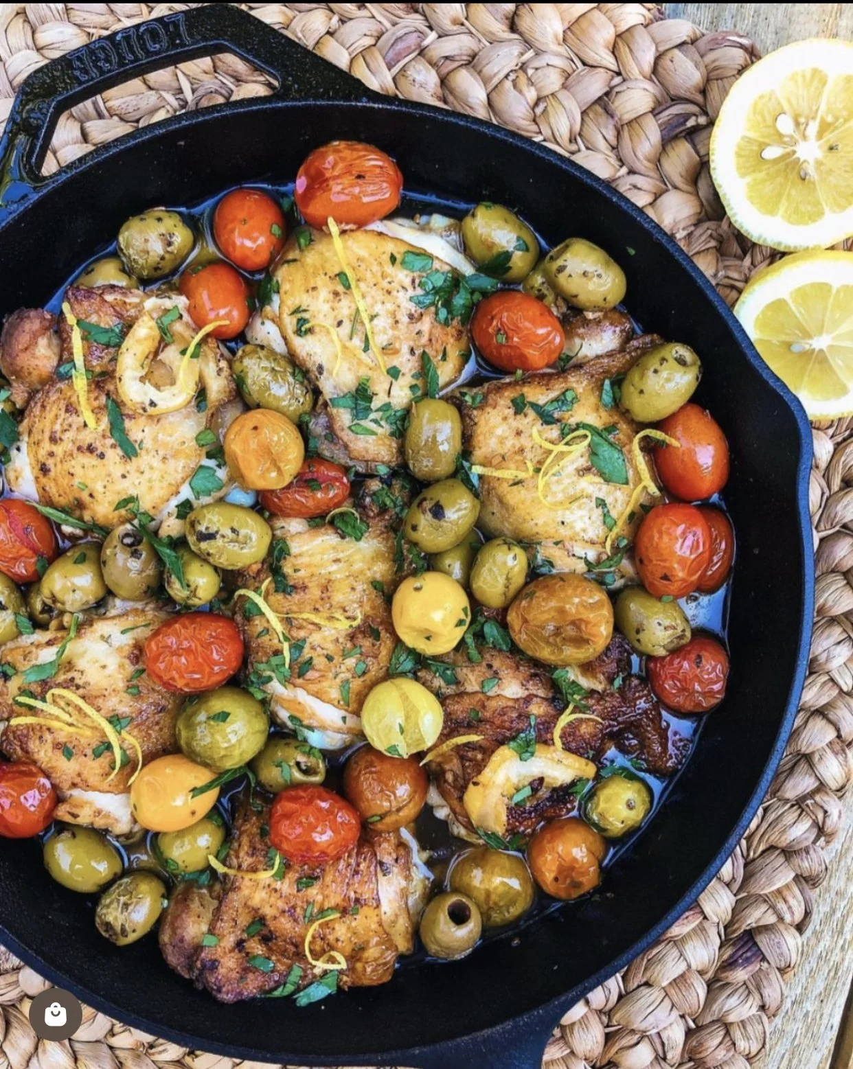 Roasted chicken thighs with cherry tomatoes and green olives garnished with chopped herbs, served in a cast iron skillet on a woven placemat with lemon slices on the side.