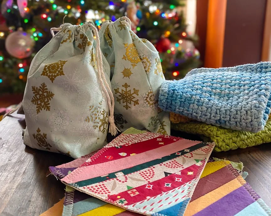Several cloth gift bags with gold snowflake and star patterns, colorful fabric napkins or tablecloths, and folded knitted or crocheted cloths on a wooden table, with a decorated Christmas tree in the background.