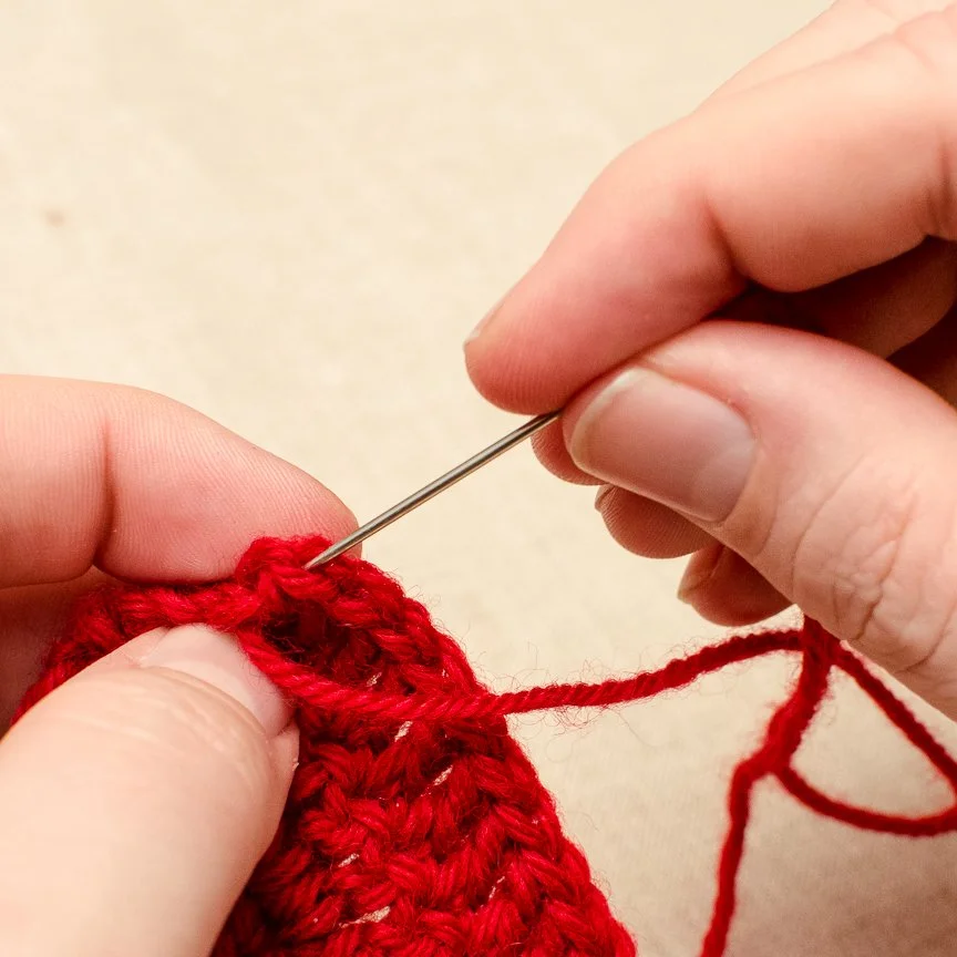 A person knitting with red yarn using a sewing needle.