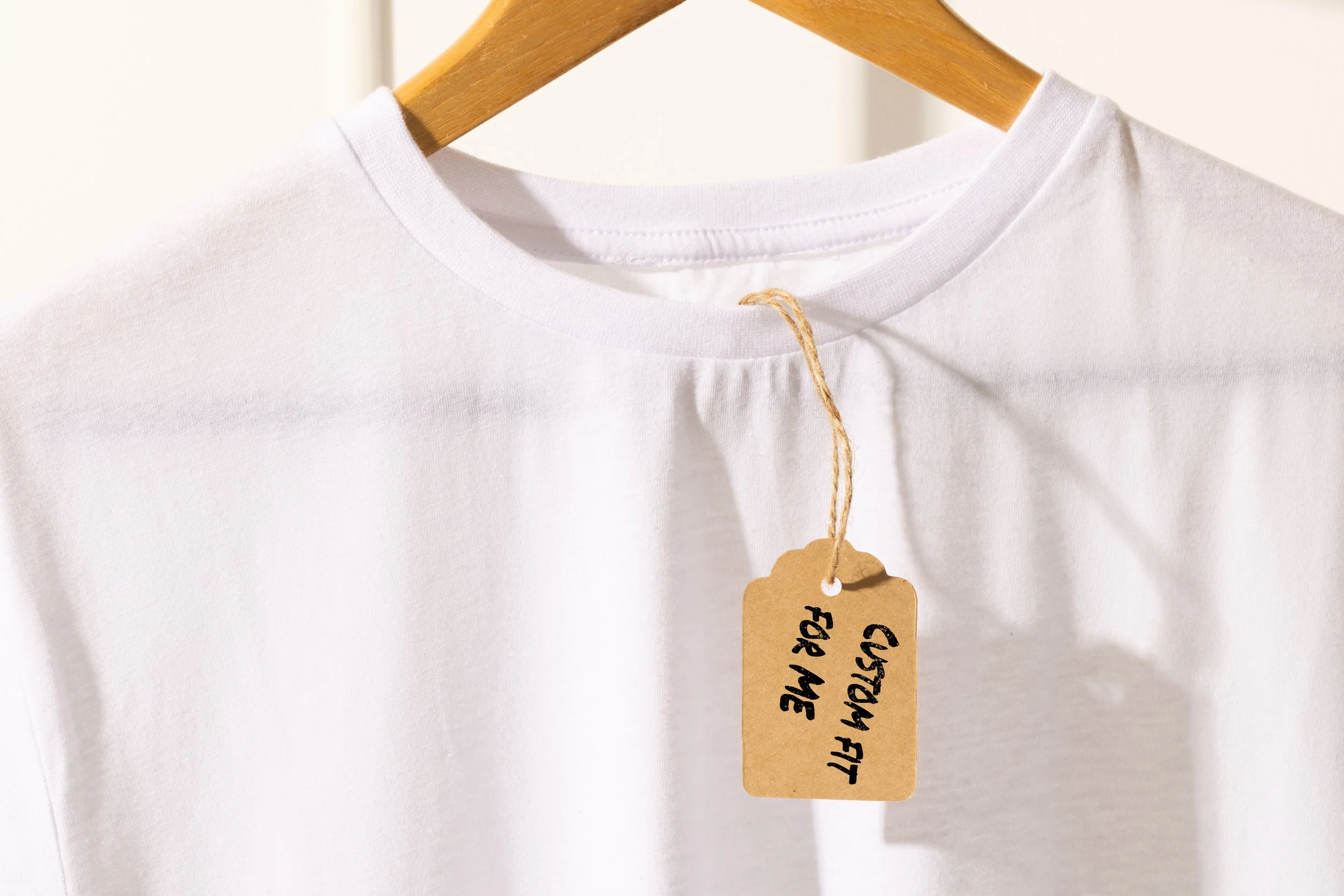White T-shirt hanging on a wooden hanger with a cardboard tag labeled "Custom Fit for Me" attached with twine