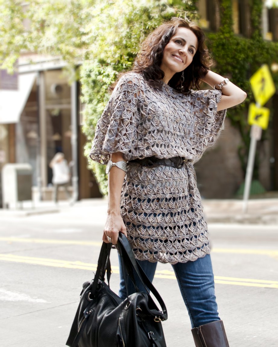 A woman with curly brown hair smiling and standing on a city street. She is wearing a crocheted beige top and jeans, carrying a large black purse.