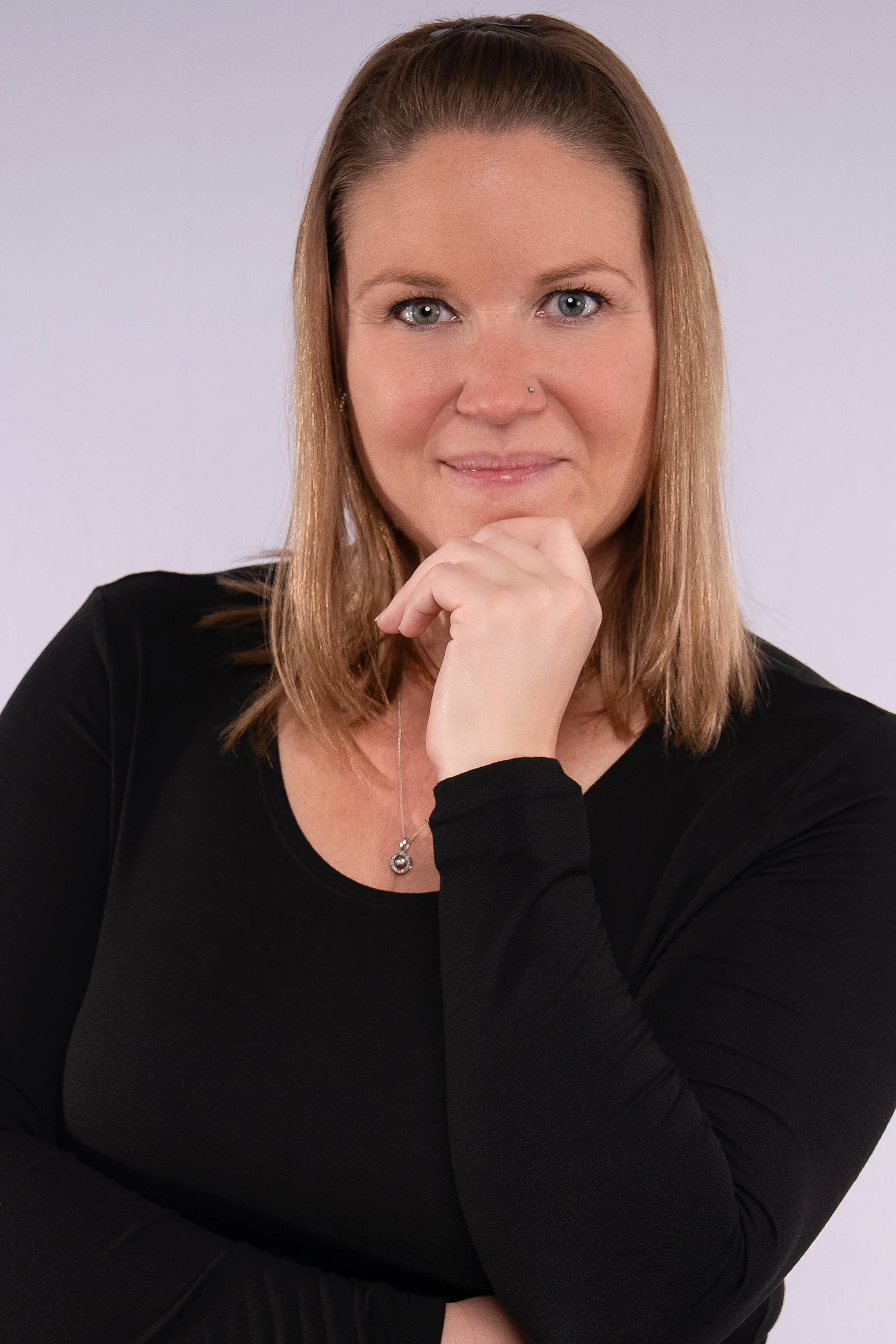 A woman with shoulder-length light brown hair, blue eyes, and a nose piercing, wearing a black top and silver necklace, is posing with her chin resting on her hand against a plain light background.