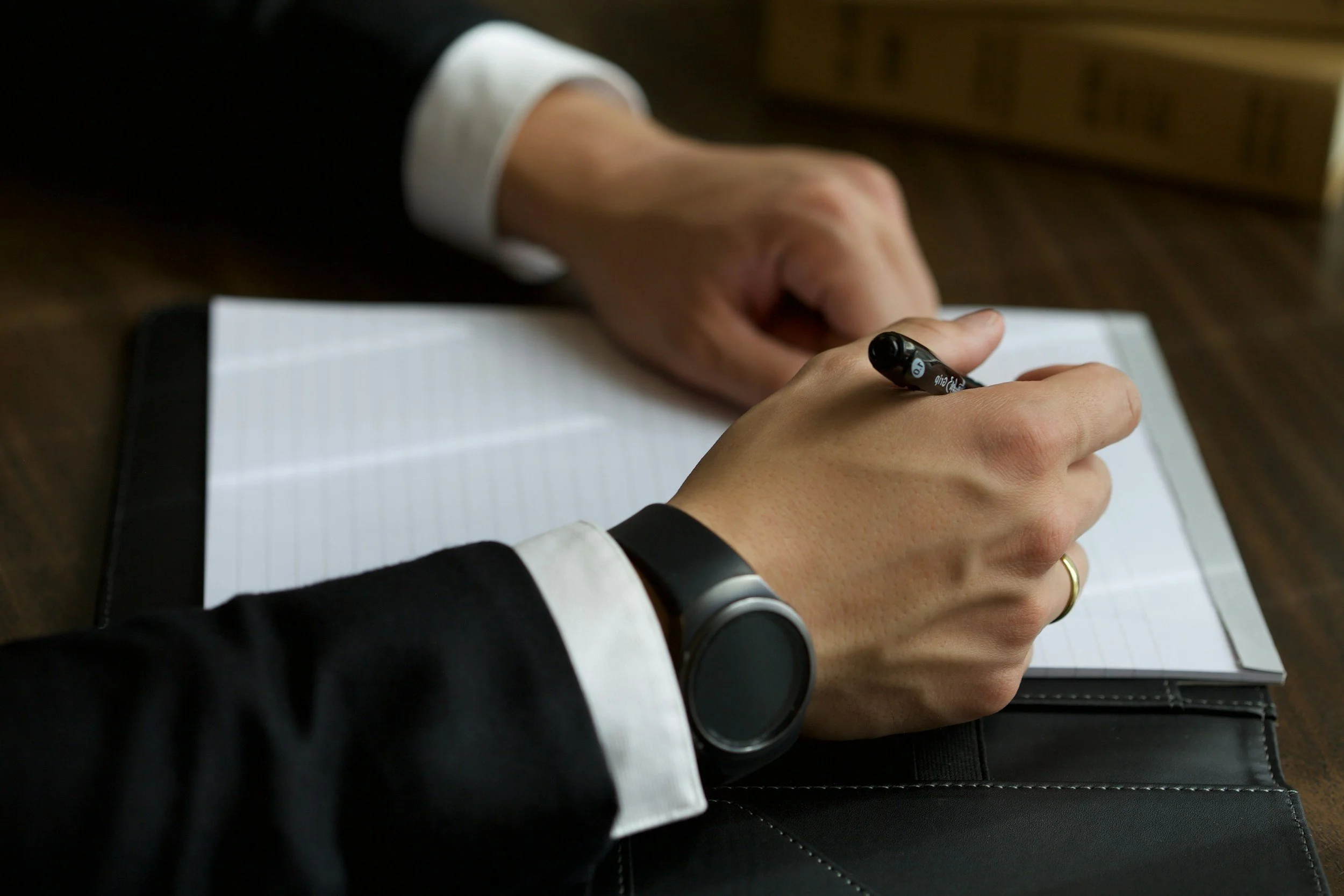Hands of a person in a business suit writing in a notebook with a black marker, wearing a smartwatch and a wedding ring, on a dark wooden desk.