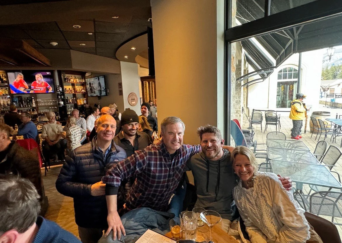 Group of five people smiling and sitting together at a bar or restaurant, with a patio area outside. The indoor setting has a bar with bottles and glasses, and sports games playing on TV screens. Outside, some chairs and a person in a yellow jacket a