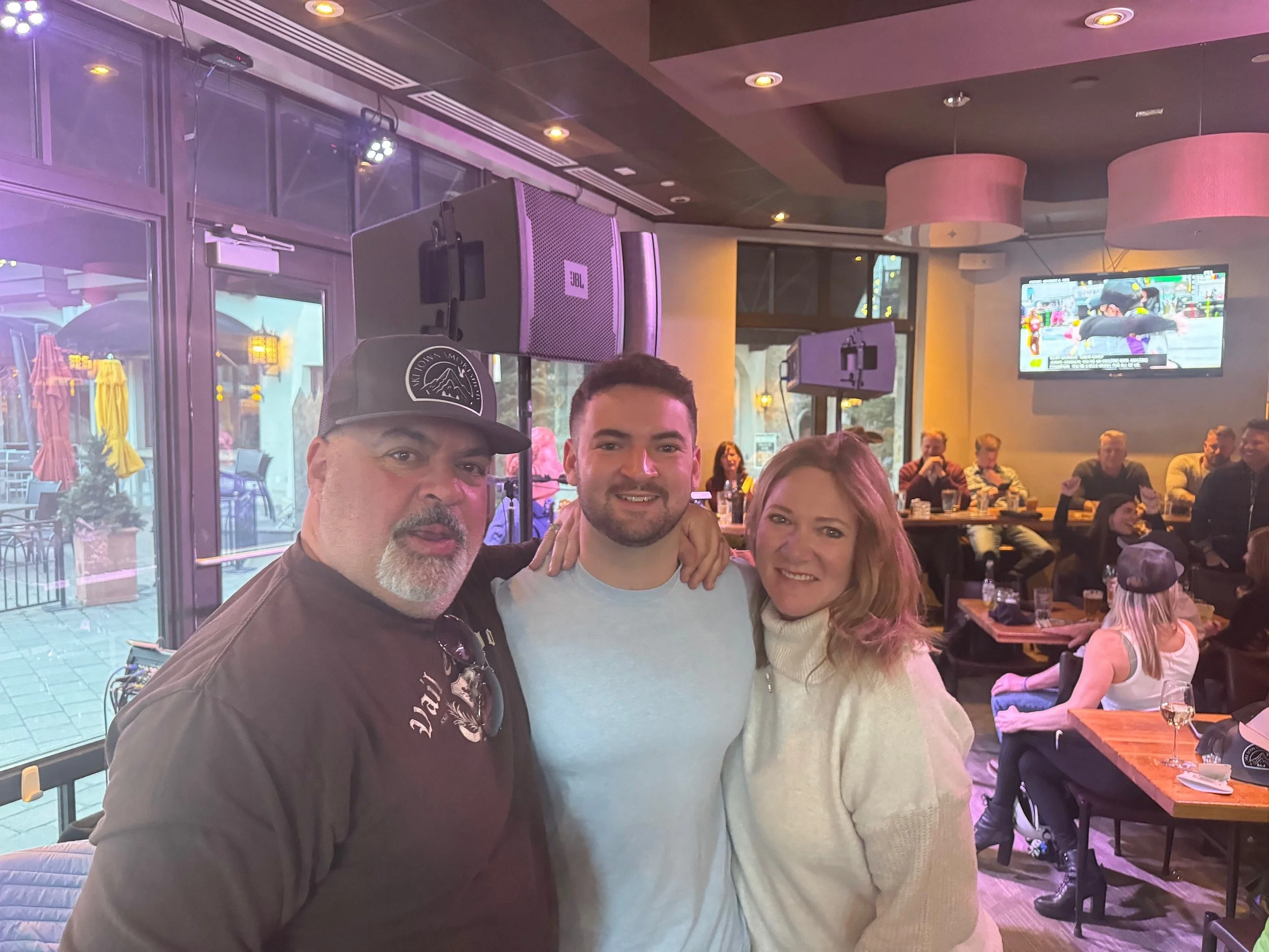 Three people smiling and posing for a photo in a lively restaurant or bar with a crowd in the background, large windows, and televisions on the wall.