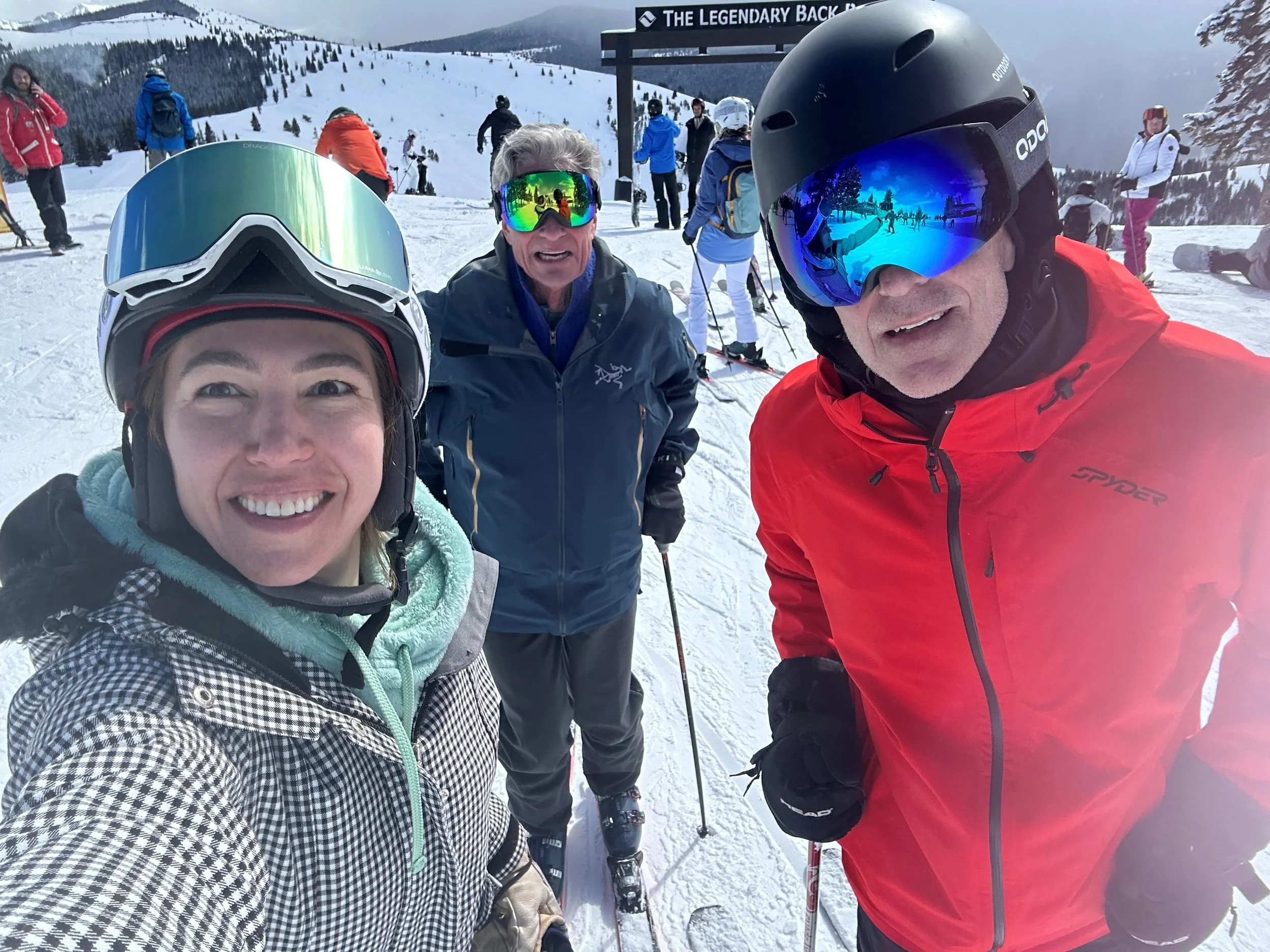 Three people in ski gear taking a selfie on a snowy mountain with other skiers and snow-covered trees in the background.