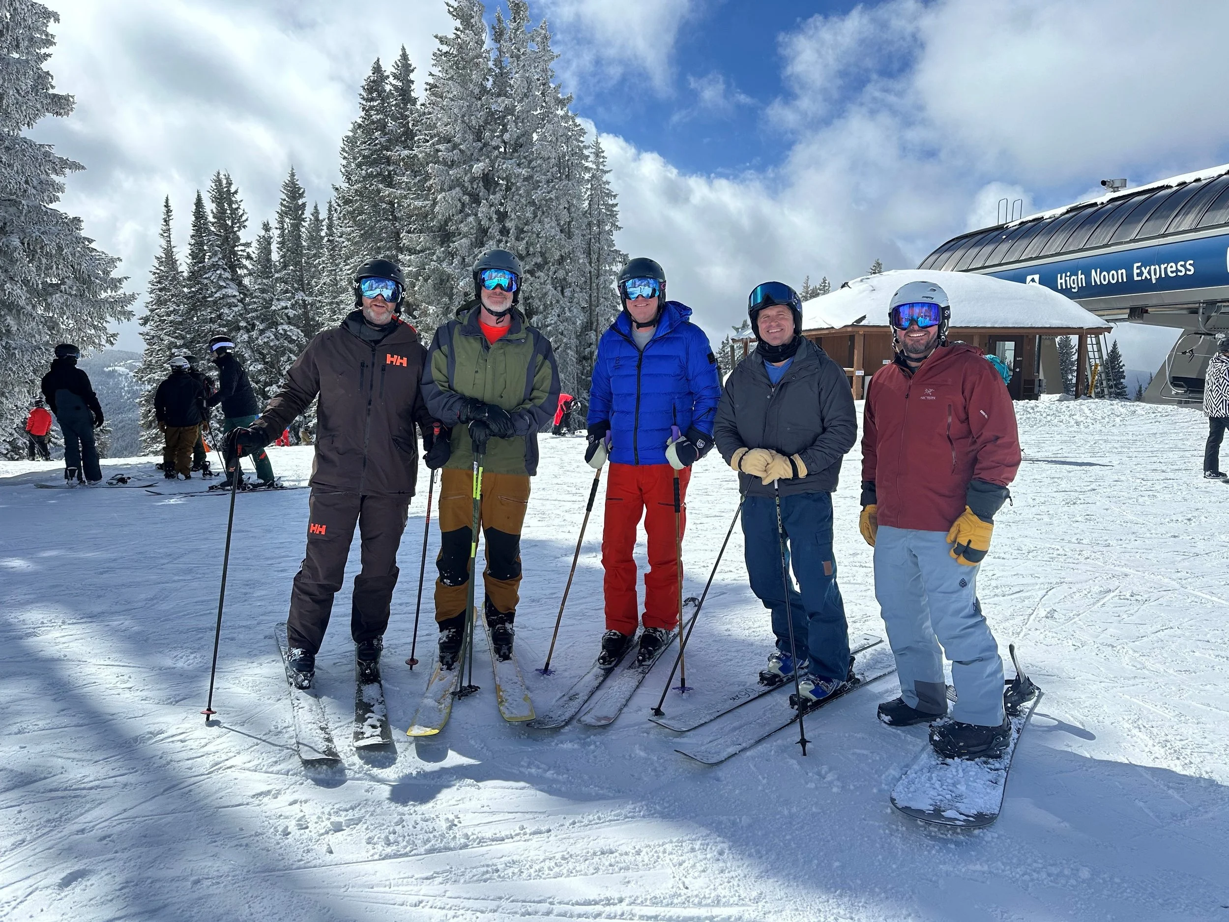 Five people in ski gear standing on snow at a ski resort, smiling and posing for the photo with skis and poles. There are snow-covered trees in the background and a building with a sign that says 'High Noon Express'.