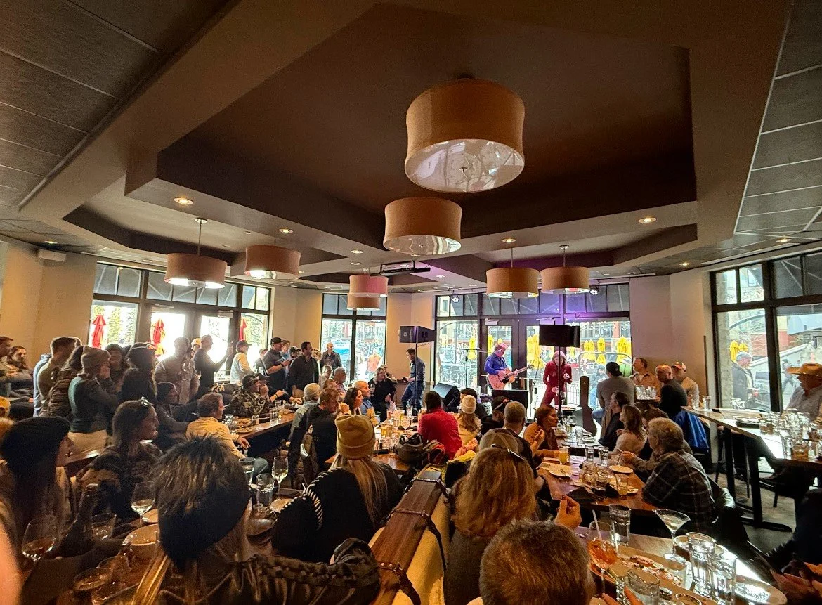 Indoor restaurant or cafe with a live musical performance, people sitting at tables, some standing and watching the performers, large windows with natural light, modern decor with pendant lighting.