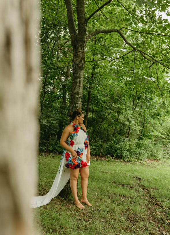 A woman in a floral dress standing barefoot in a lush green wooded area with trees and grass, partially obscured by a blurred tree trunk in the foreground.
